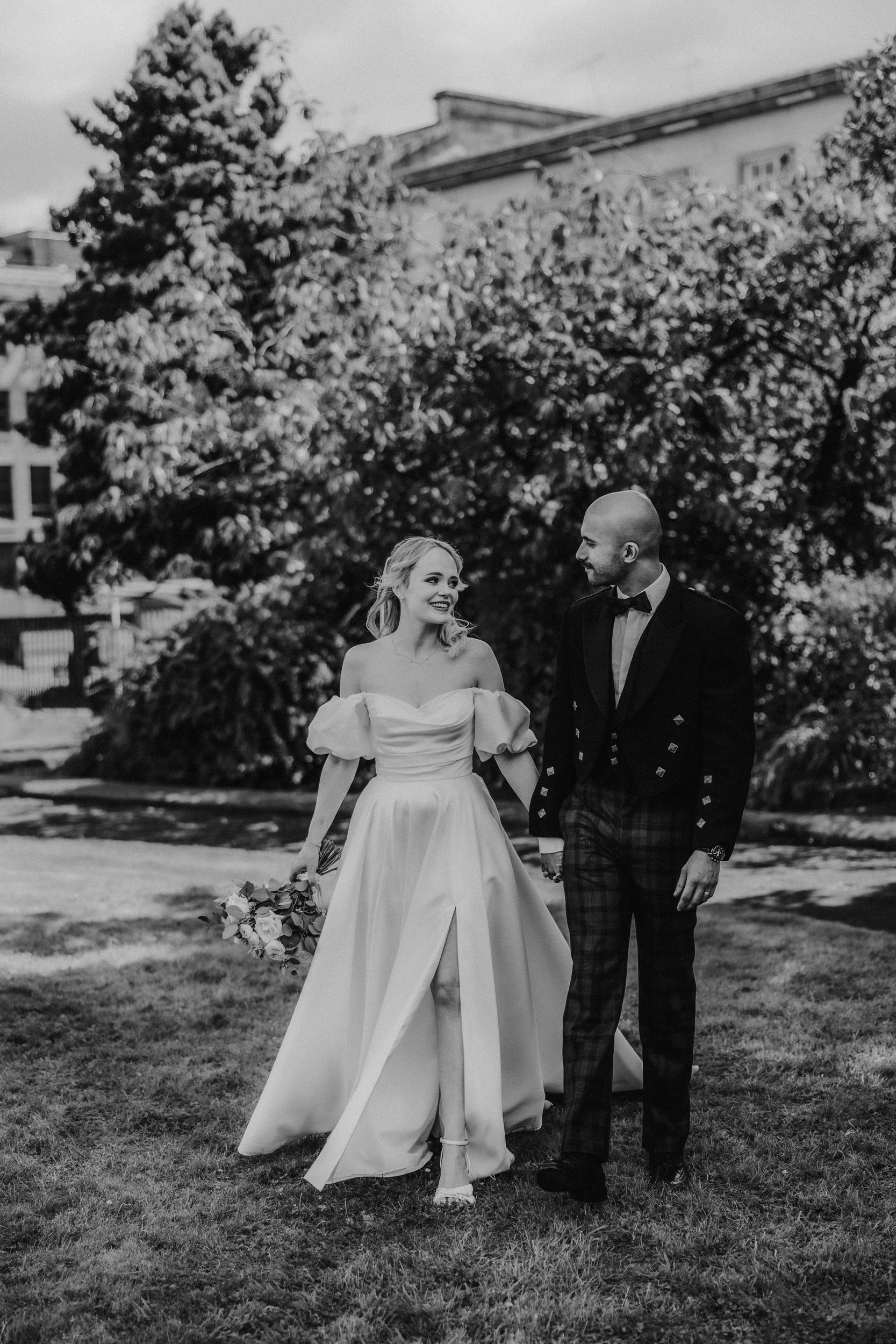 Bride in off-shoulder gown holding a bouquet and groom in jacket and tartan trousers walking hand in hand outdoors.