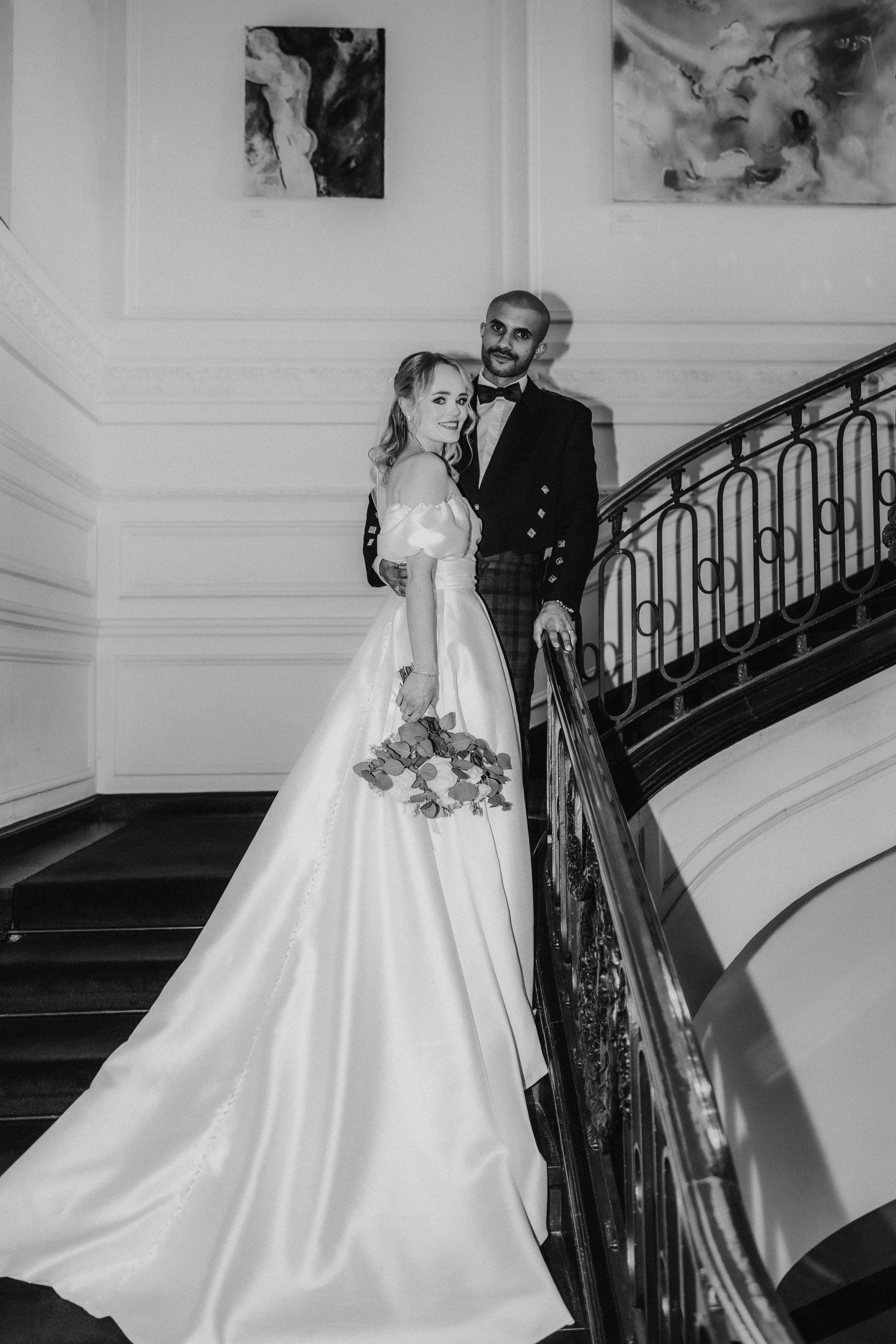 Bride in off-shoulder satin gown with bouquet and groom in formal jacket and tartan kilt on a sweeping staircase.