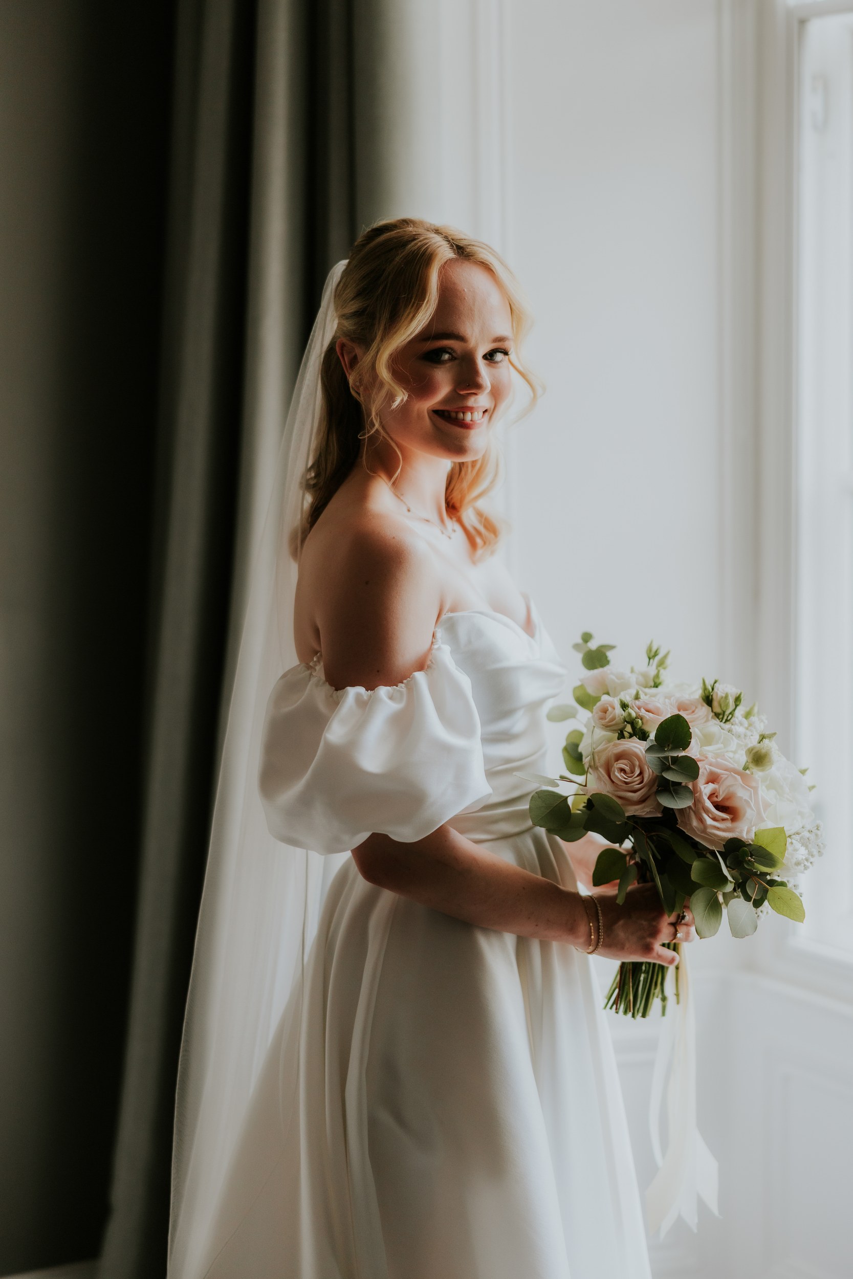 Bride in off-shoulder wedding dress and veil holding a pale rose bouquet, smiling by a bright window.