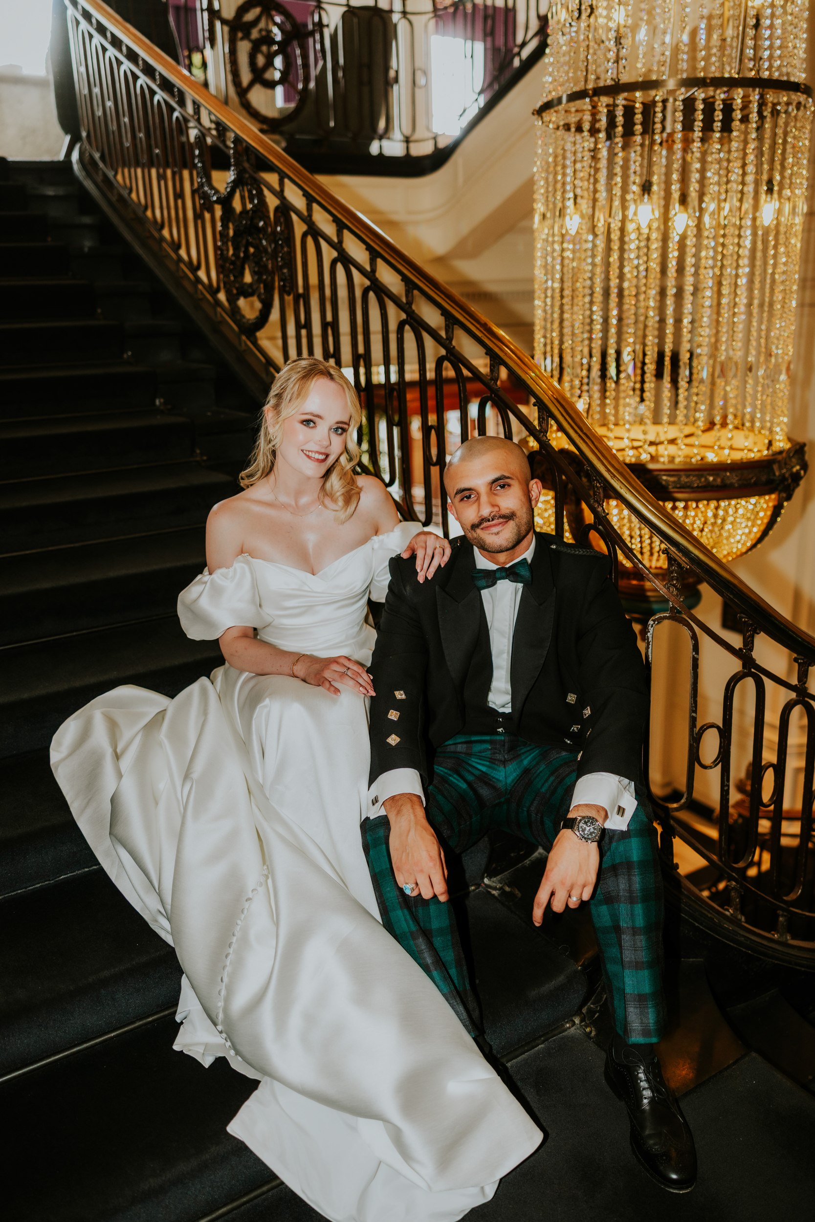 Bride in off-shoulder white gown and groom in jacket with tartan trousers seated on a hotel staircase beside a crystal chandelier.