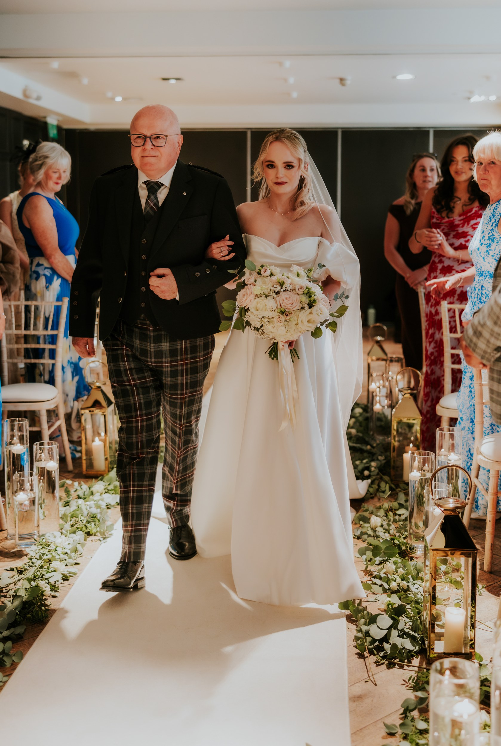 Bride in off-shoulder white gown and veil walks down a candlelit aisle arm-in-arm with an older man, holding a cream and blush bouquet.