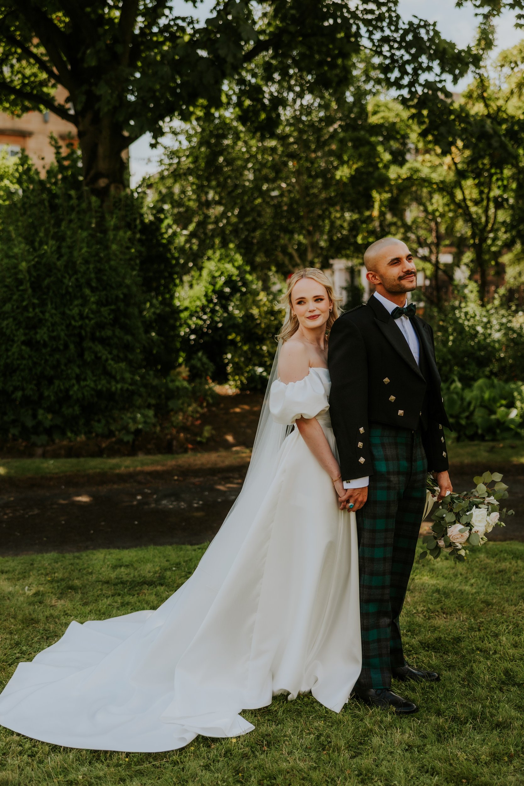 Bride in off-shoulder white gown leans against groom in tartan kilt and dark jacket, holding a bouquet in a leafy garden.