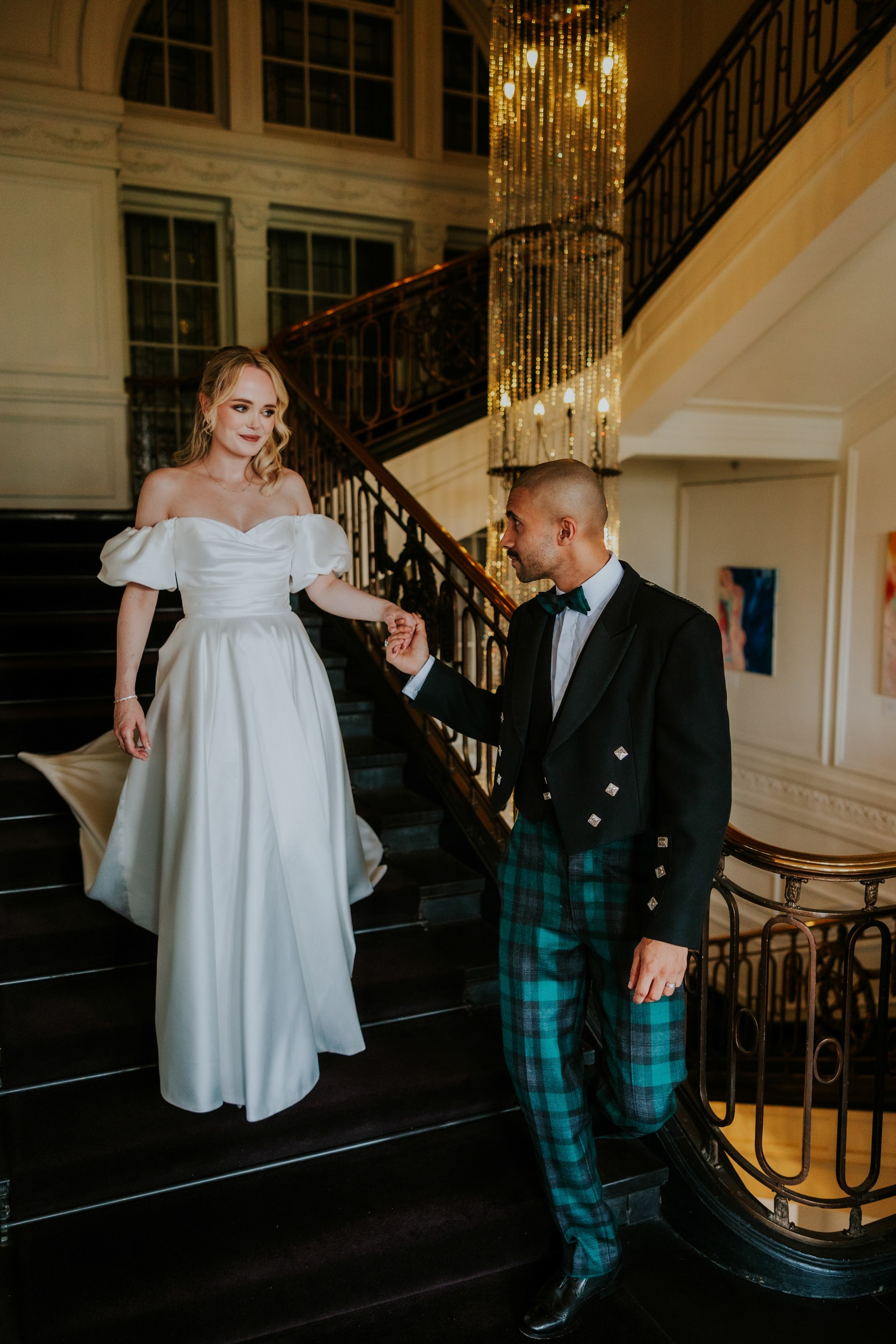 Bride in off-the-shoulder gown and groom in tartan trousers holding hands on a grand hotel staircase.