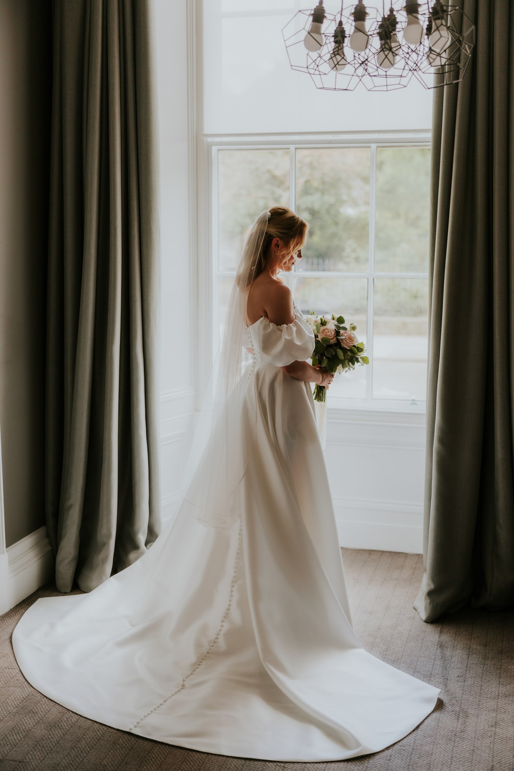 Bride in off-the-shoulder gown and veil holding bouquet by a large window with grey curtains.