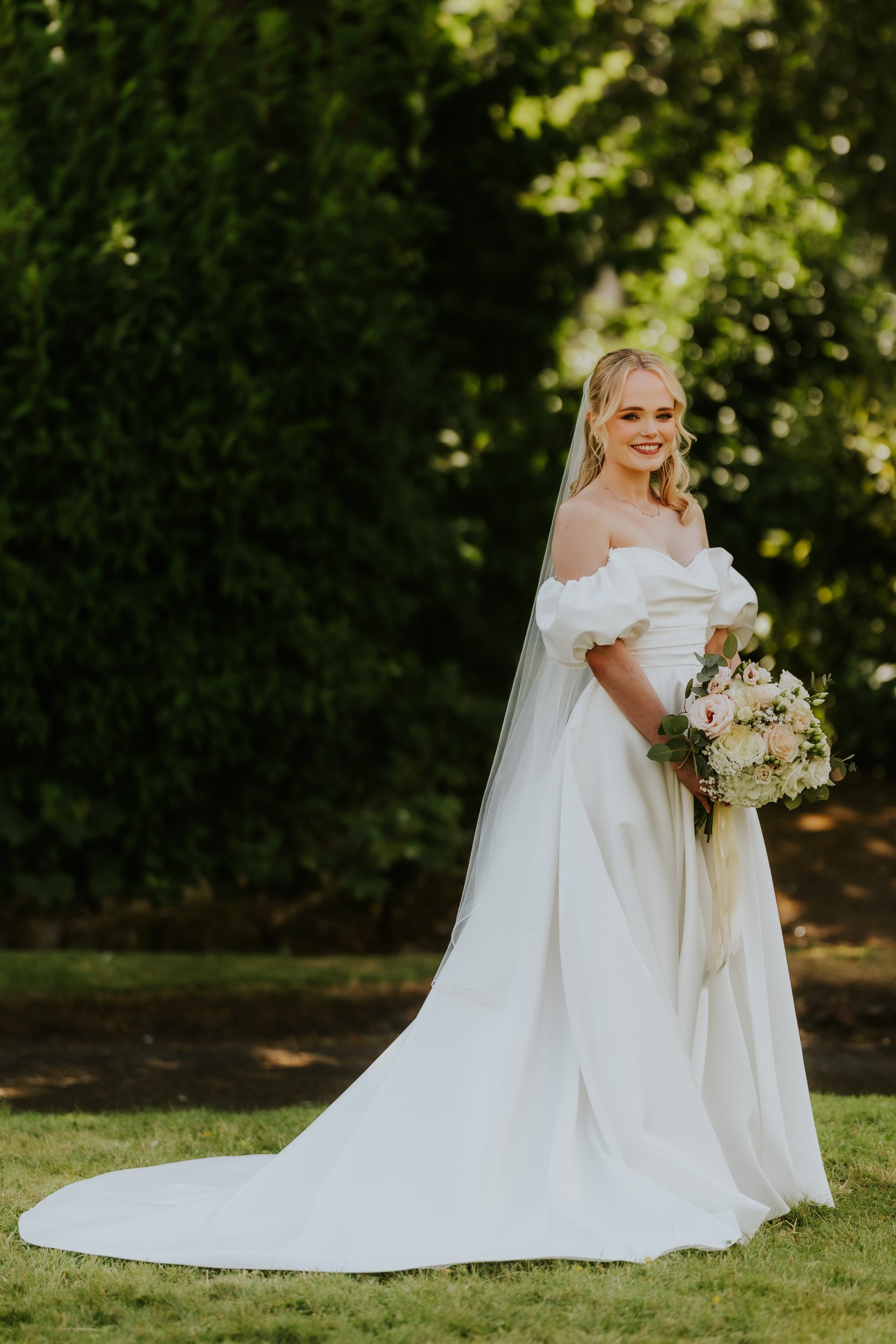 Bride in off-the-shoulder gown and veil holding a pale rose bouquet, standing outdoors against leafy greenery.
