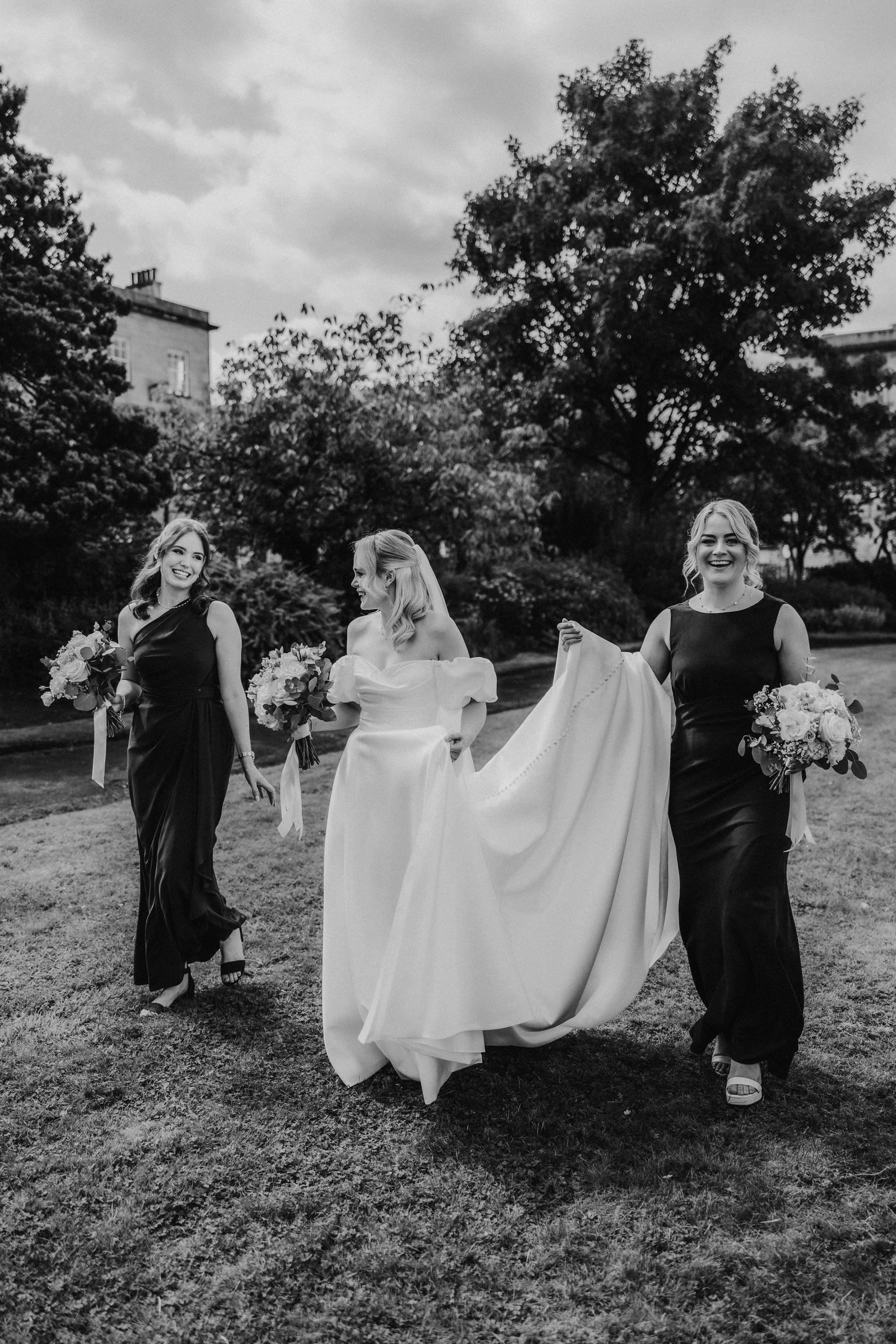 Bride in off-the-shoulder gown walking with two bridesmaids holding bouquets, one lifting her train on a lawn.
