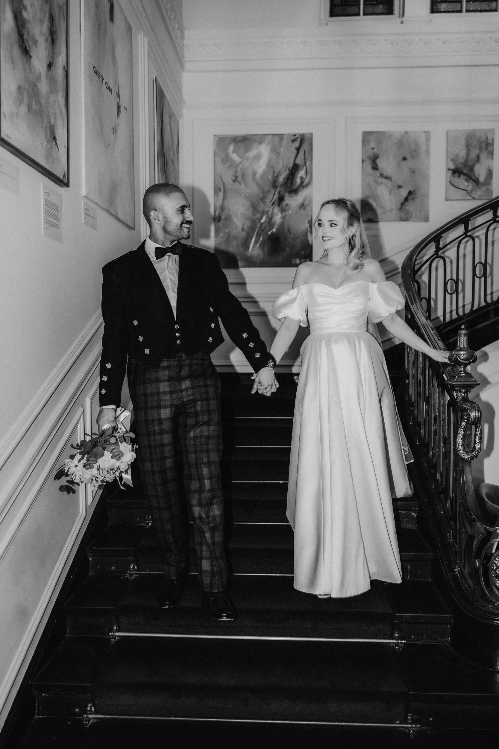 Bride in off-the-shoulder satin gown and groom in tartan kilt hold hands walking down an indoor staircase.
