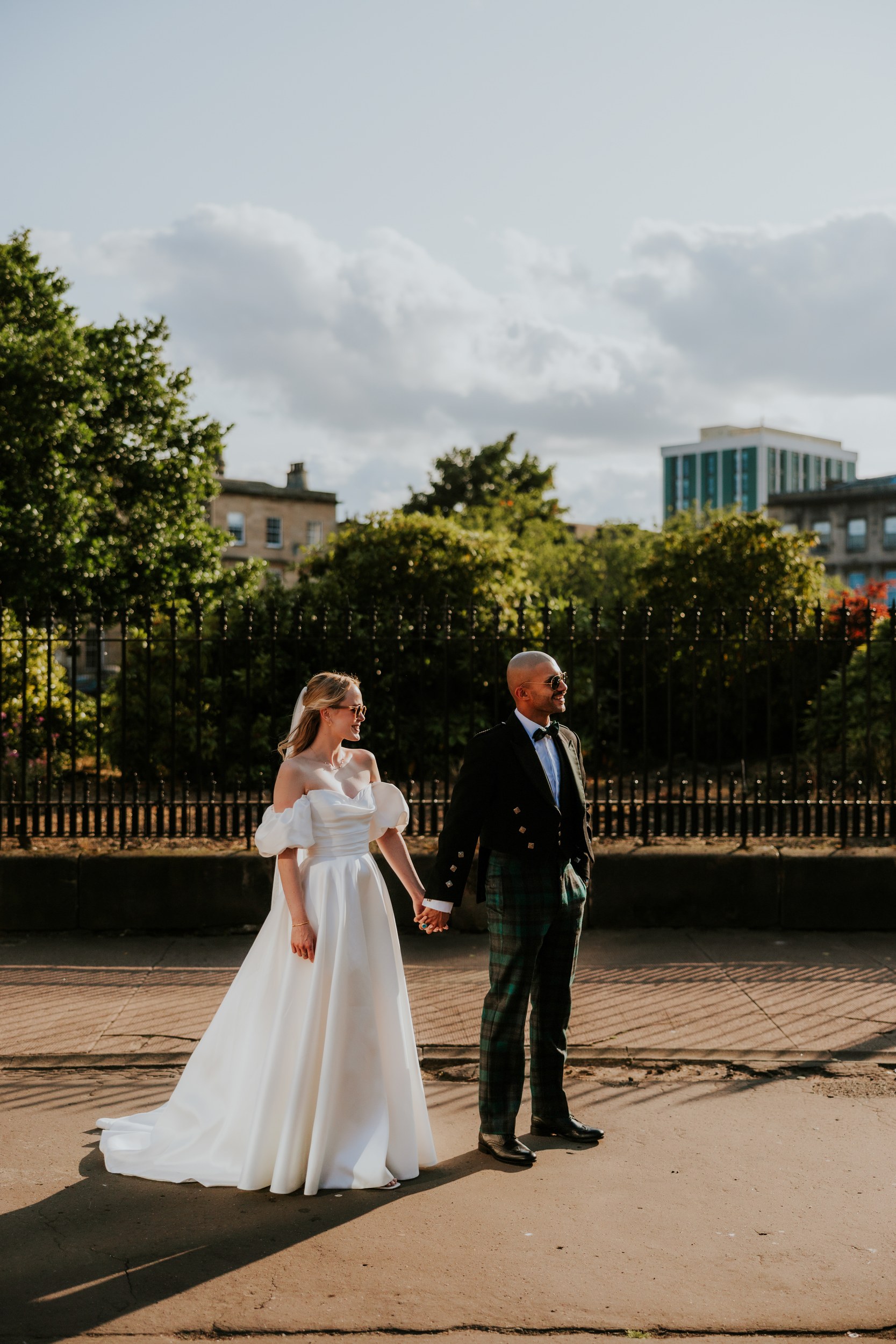 Bride in off-the-shoulder white dress and groom in kilt holding hands walking along a pavement.