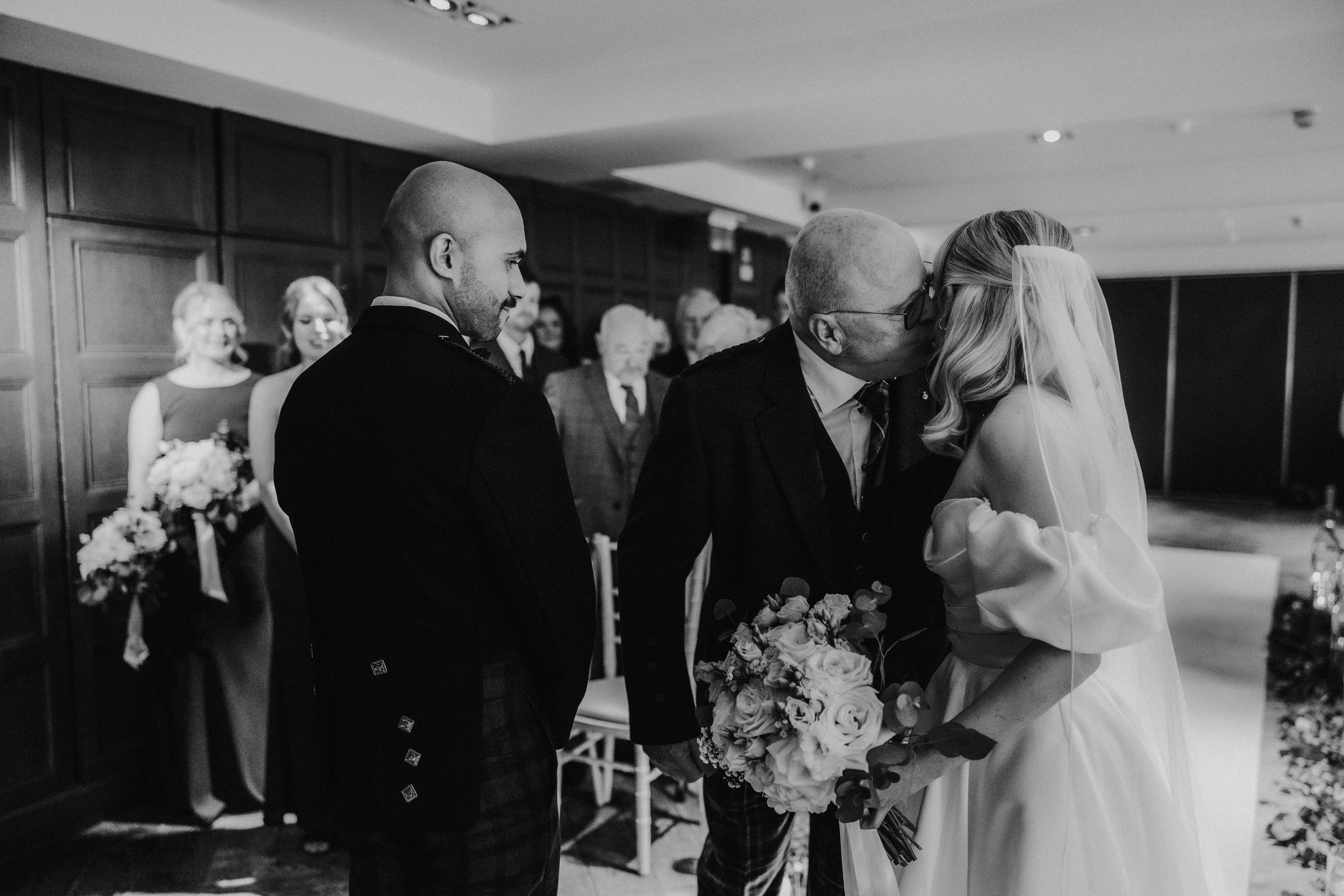 Bride in veil kissed on the cheek by an older man while the groom watches, bridesmaids and guests visible behind.