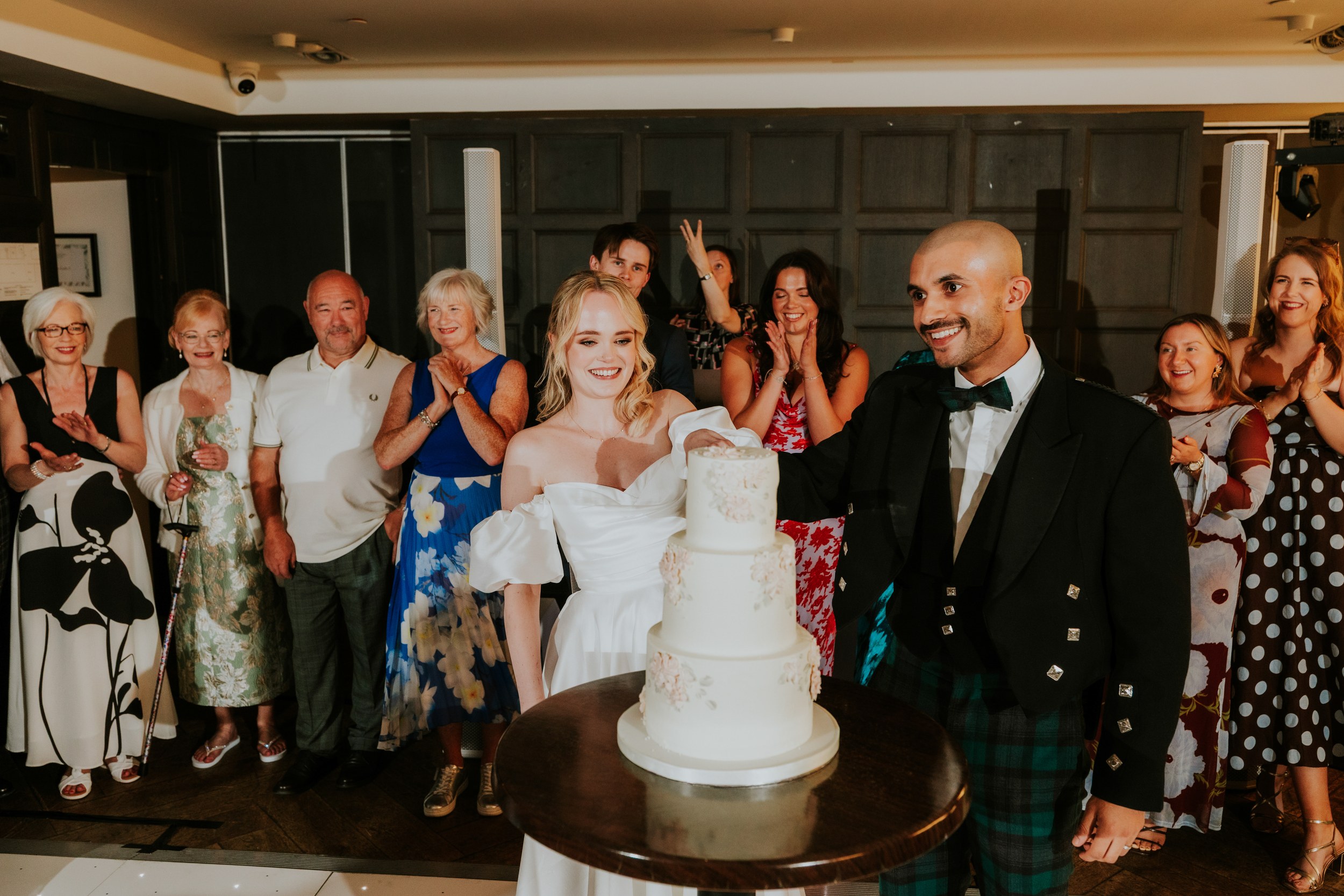Bride in white dress and groom in a kilt cut a tiered wedding cake as guests applaud.