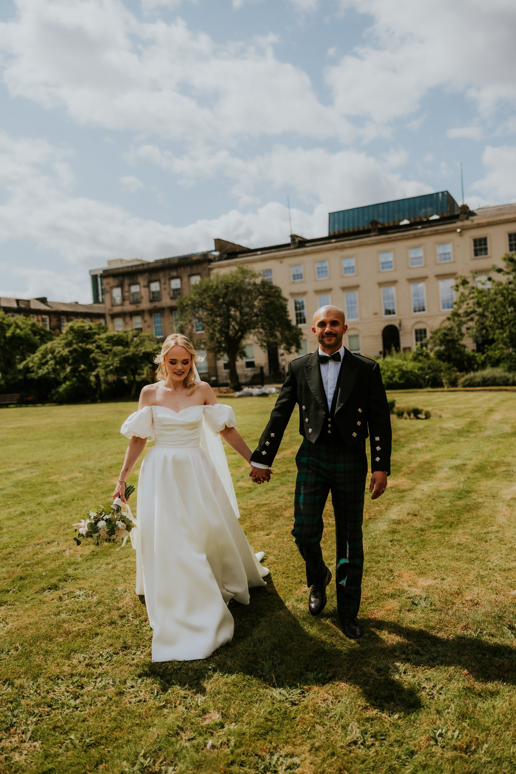 Bride in white gown and groom in formal jacket and tartan trousers holding hands on a lawn outside Kimpton Blythswood Square.
