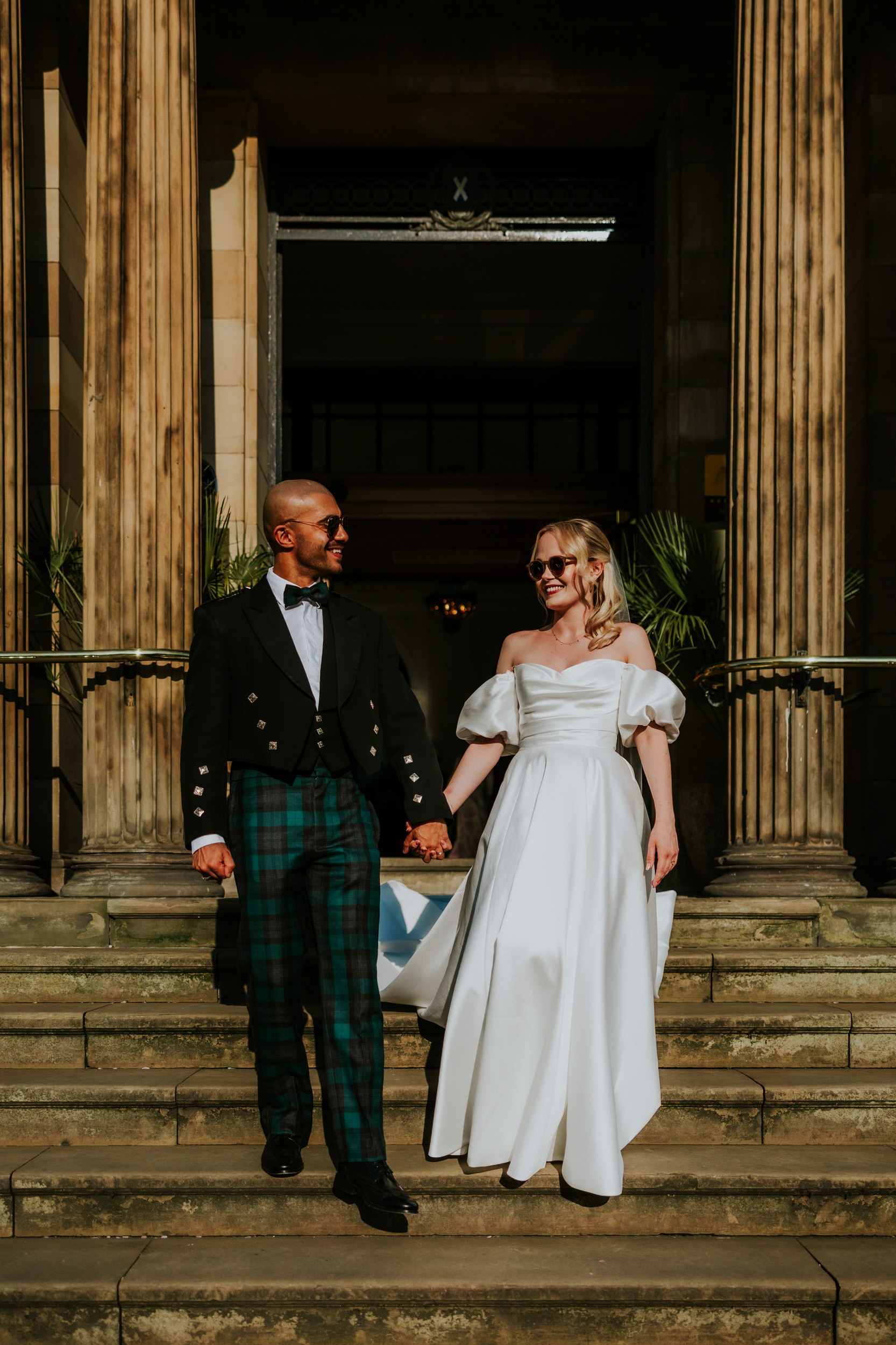 Bride in white gown and groom in tartan kilt hold hands on stone steps beneath grand columns, both wearing sunglasses.