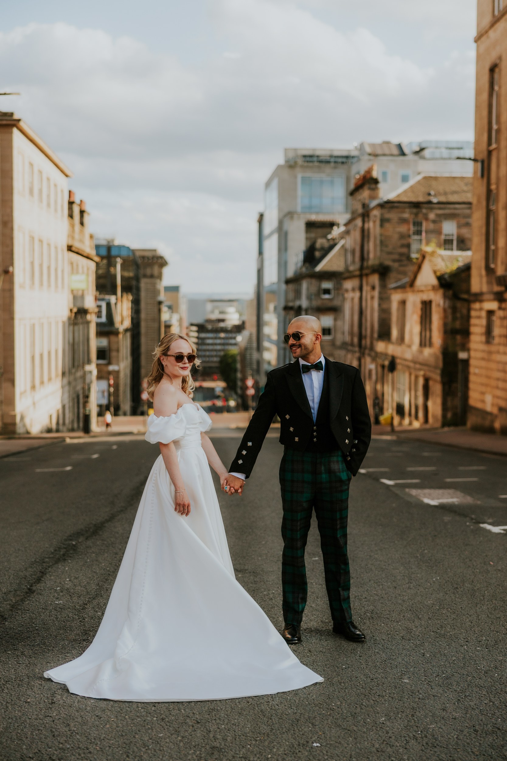 Bride in white gown and groom in tartan trousers holding hands on a city street, both wearing sunglasses.