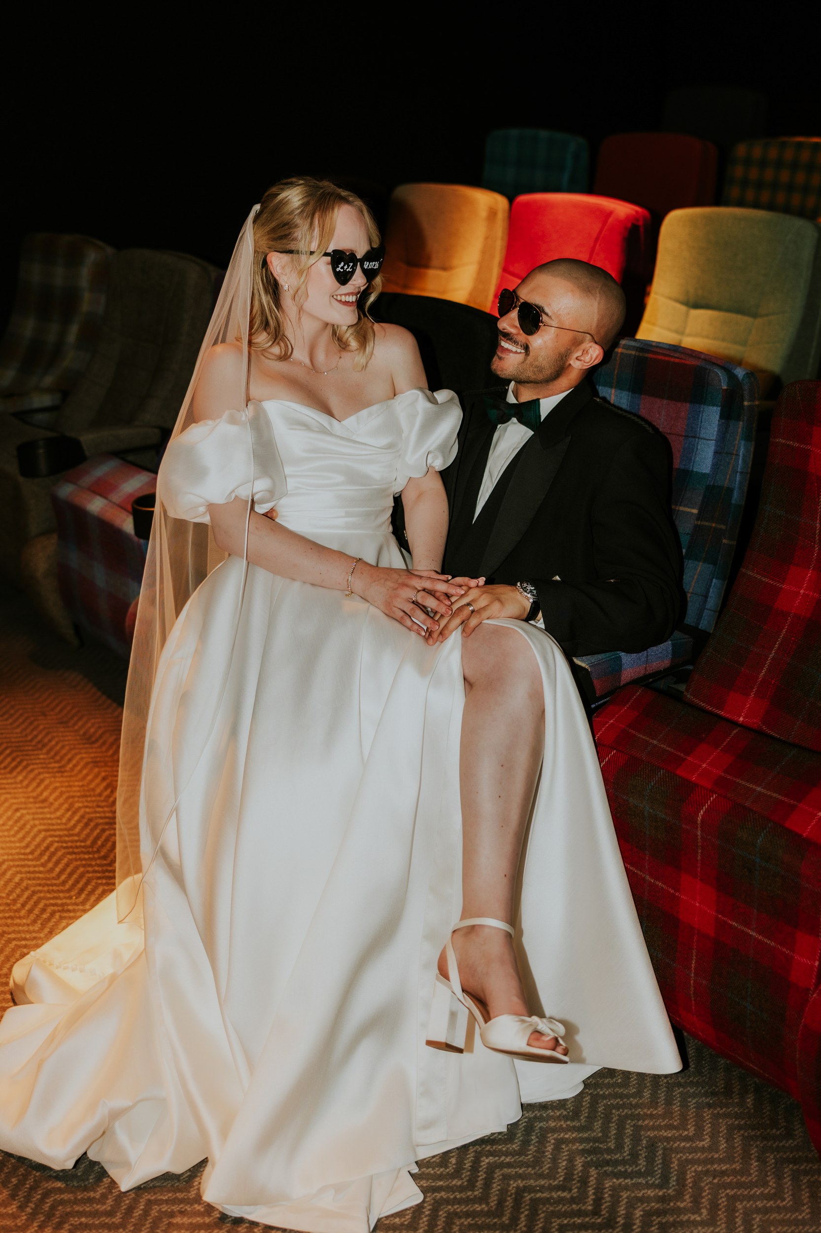 Bride in white gown and veil and groom in tux wearing sunglasses, seated in colourful theatre-style chairs holding hands.