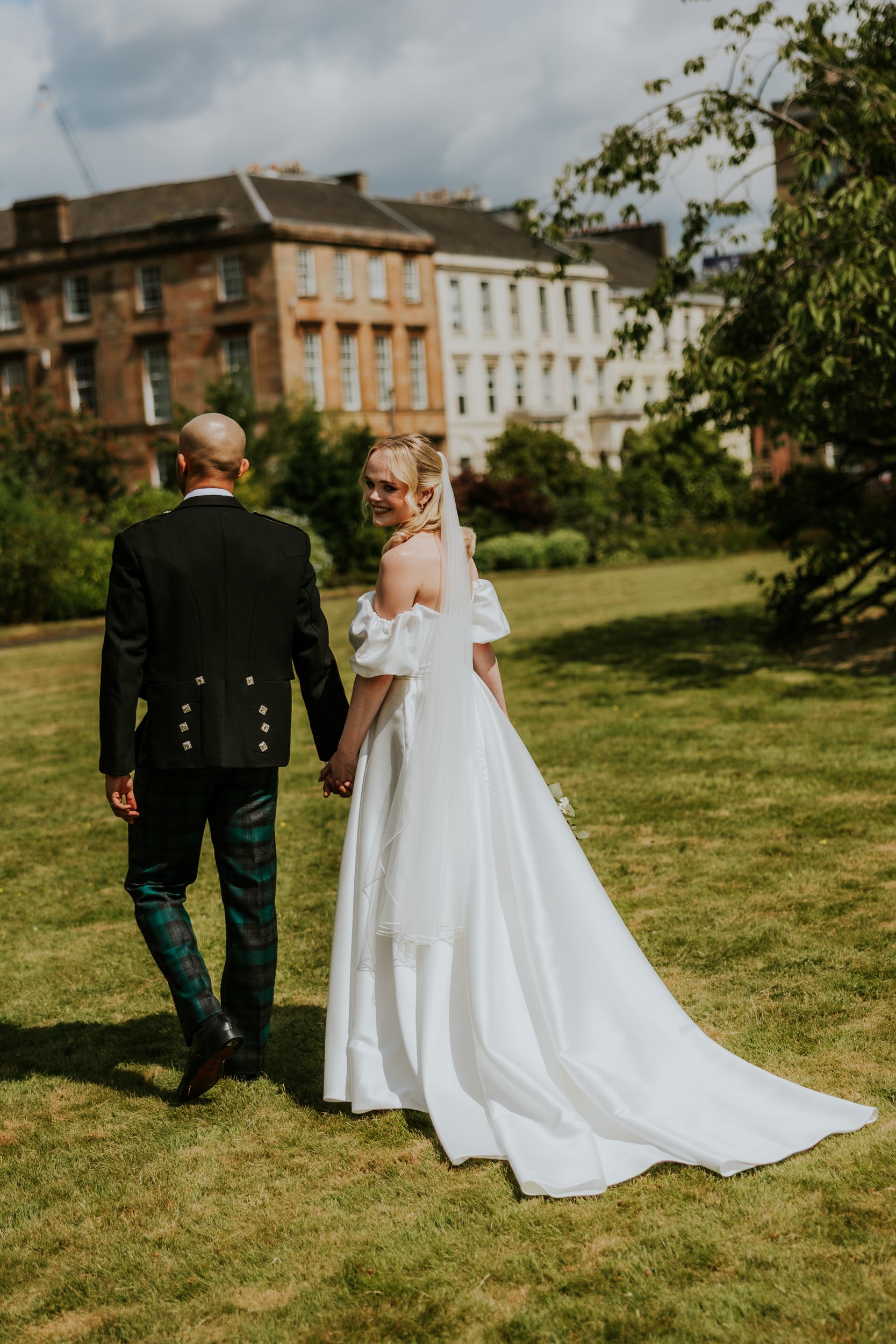 Bride in white gown and veil holding hands with groom in dark jacket and tartan trousers on a lawn, bride smiling back.
