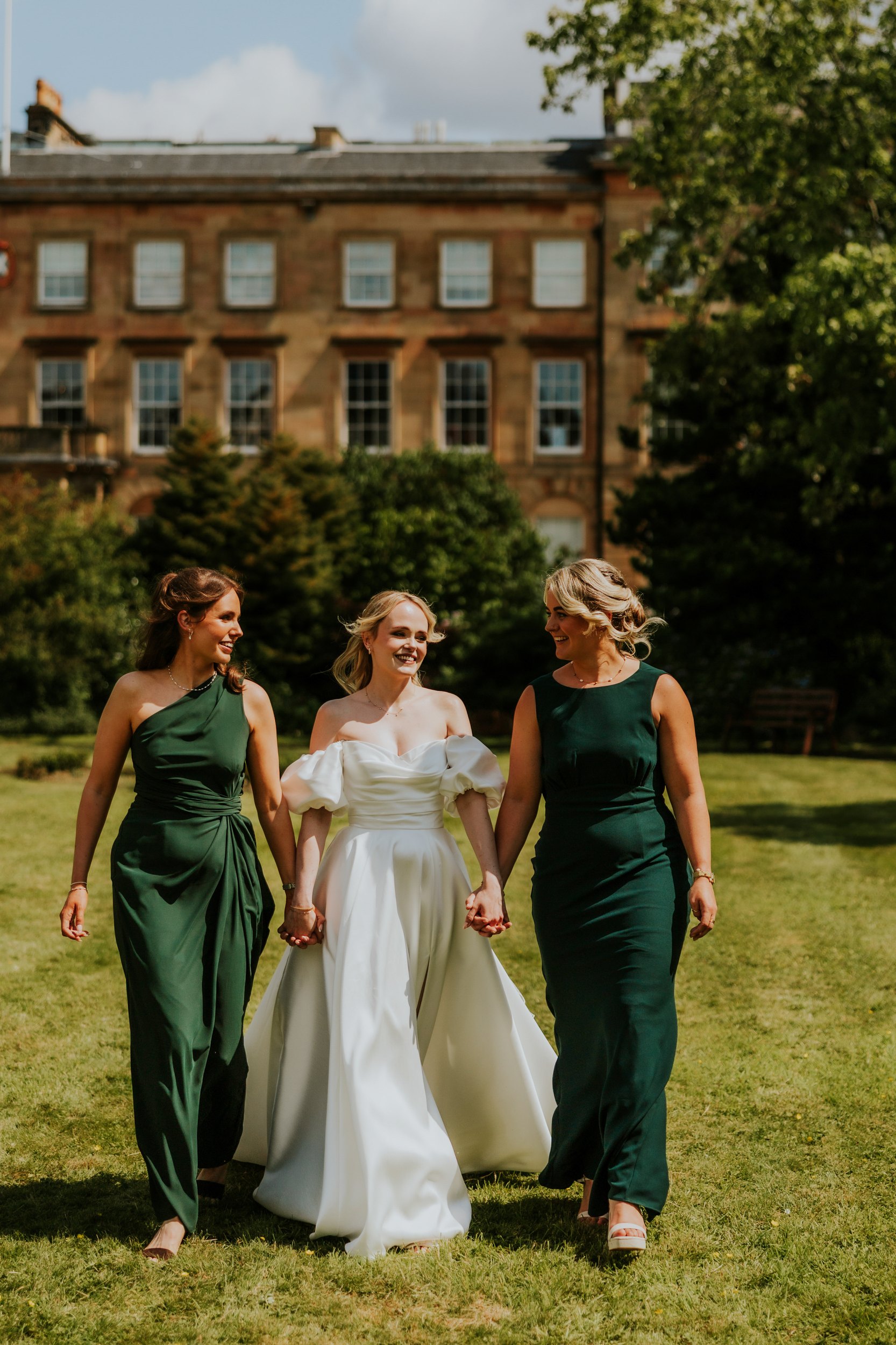 Bride in white holding hands with two bridesmaids in dark green dresses walking on a hotel lawn.