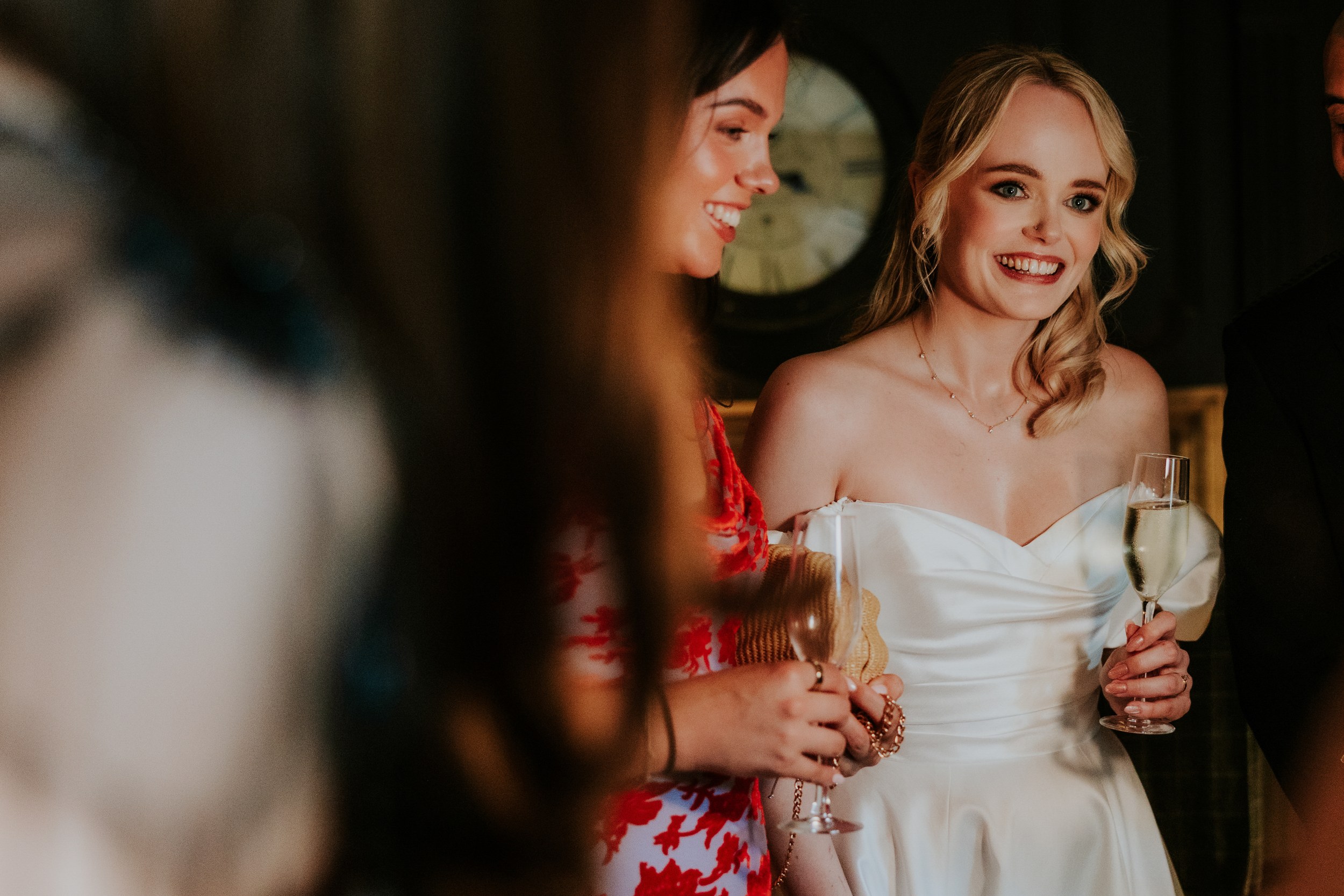 Bride in white off-shoulder dress smiling and holding a champagne glass beside a woman in a red floral dress.
