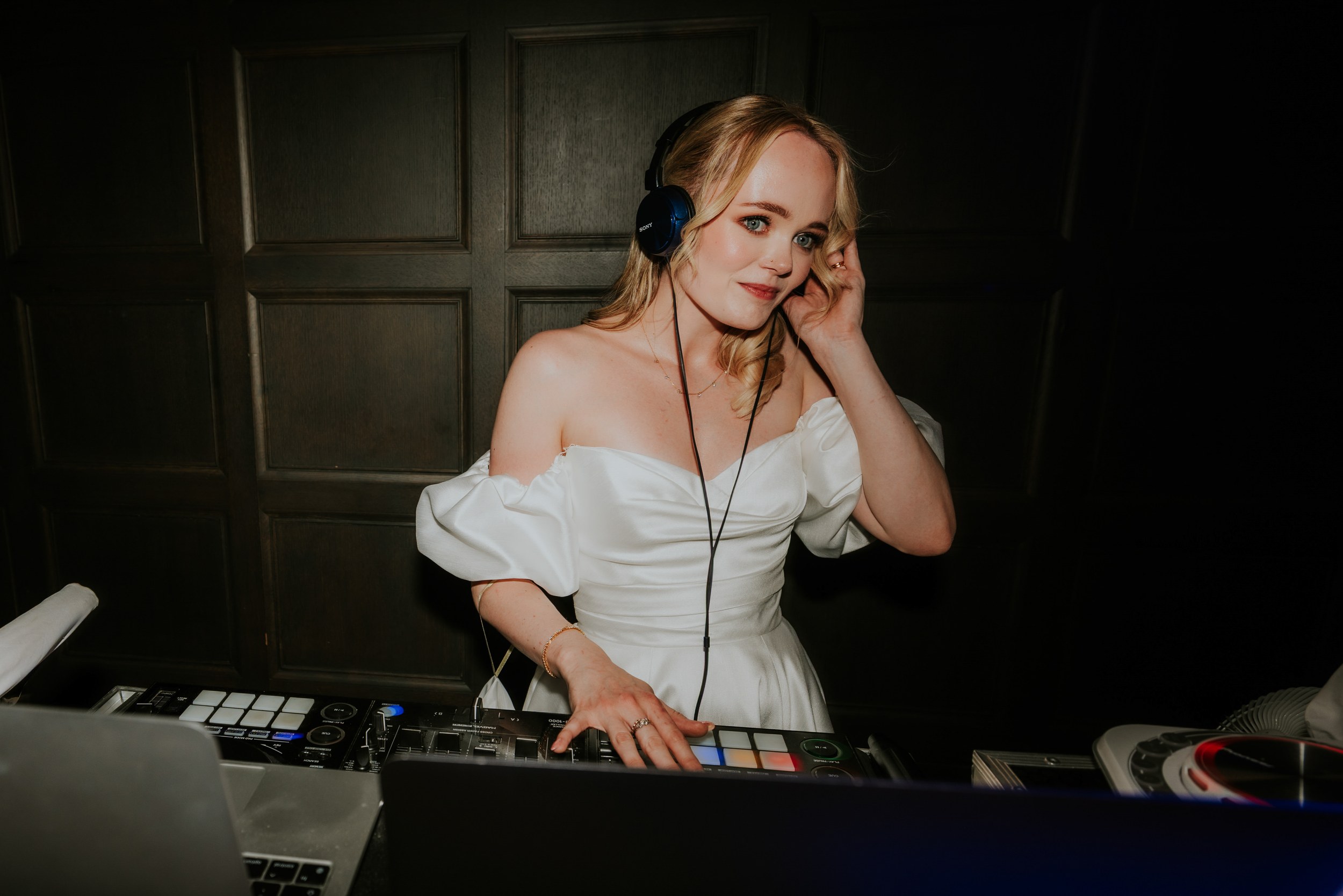 Bride in white off-the-shoulder dress wearing headphones and DJing at a mixer against a dark wood-panel wall.