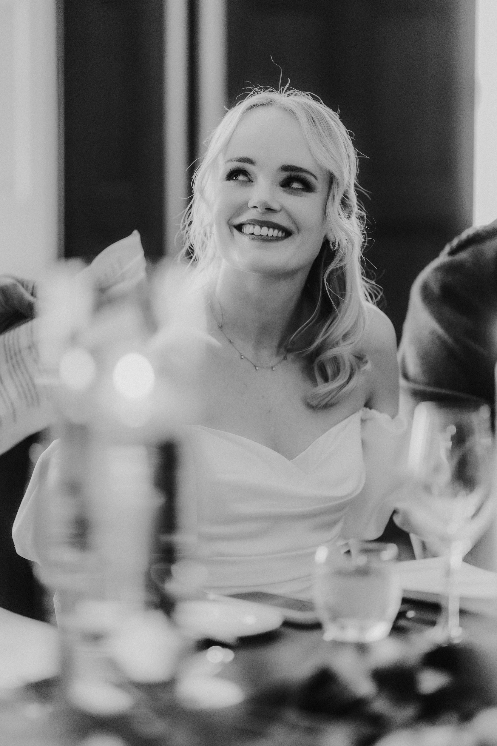 Bride smiling and looking up during the reception, close-up portrait with blurred glasses in the foreground.