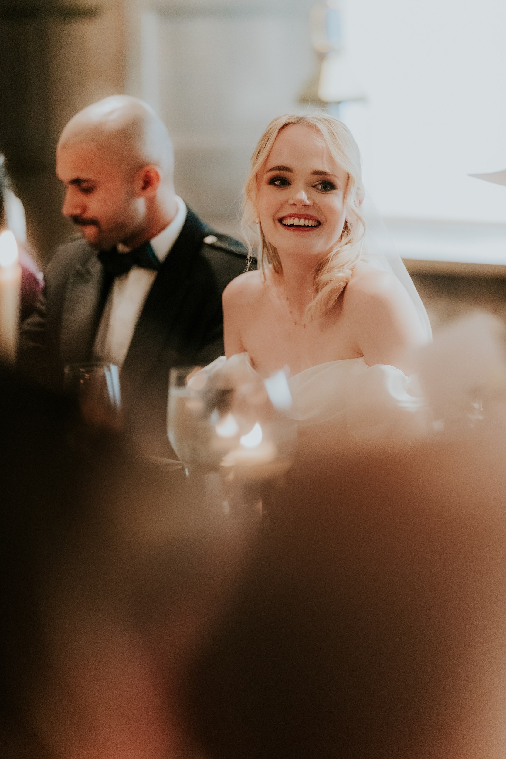 Bride smiling at a reception table beside the groom, with candles and glasses in the foreground.
