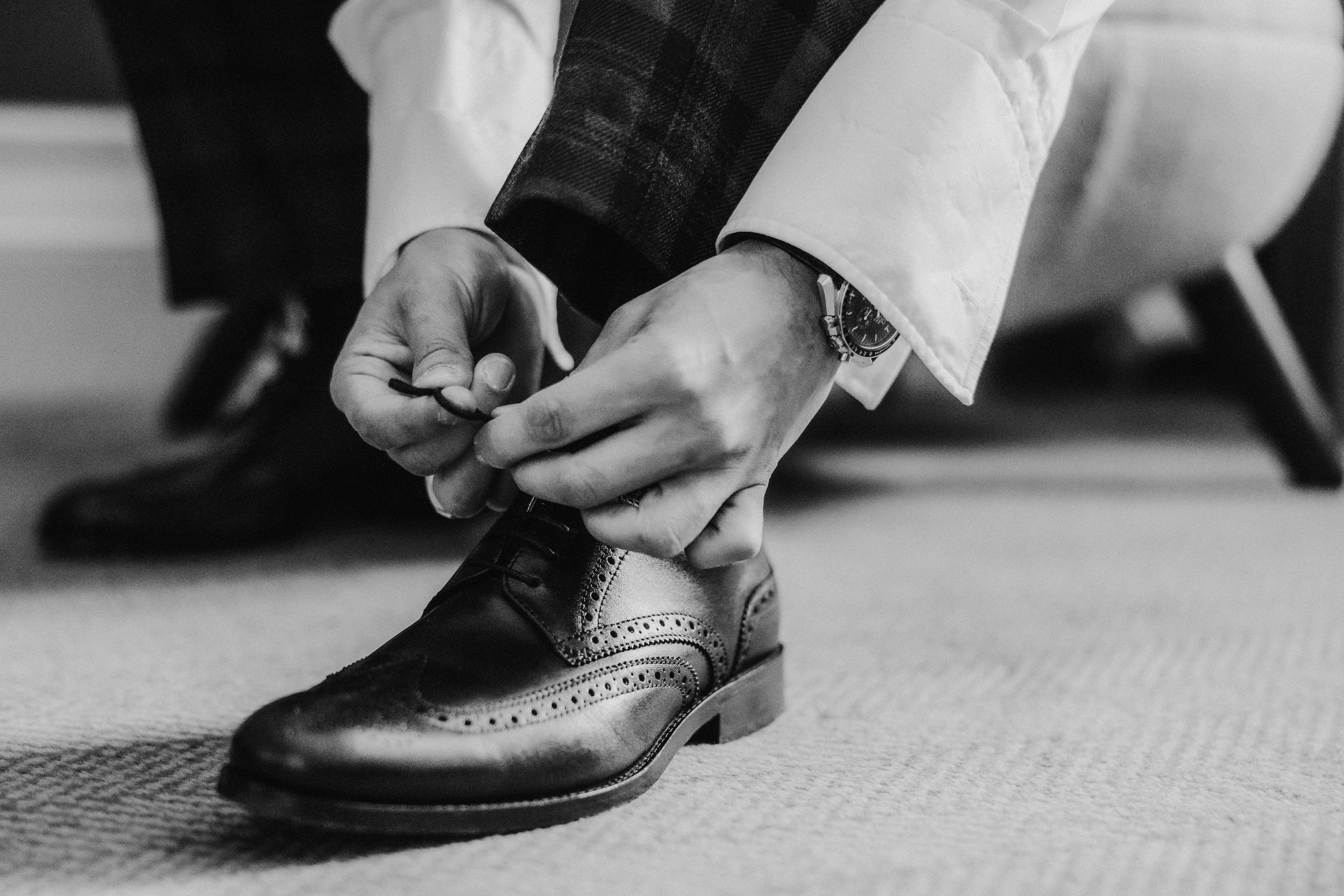 Close-up black-and-white of hands tying a leather brogue, white shirt cuff and watch visible.