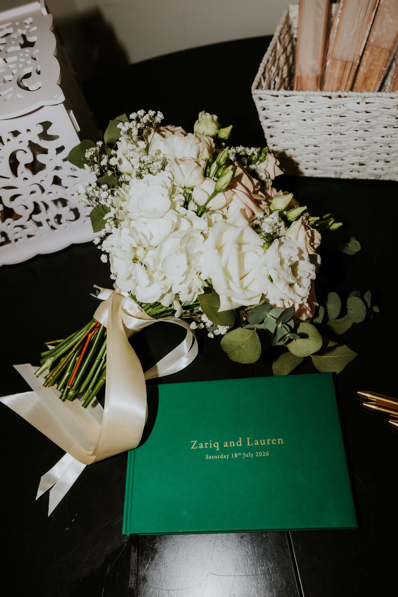 Cream and white bridal bouquet with ribbon beside a green wedding album reading 'Zariq and Lauren 18 July 2026'.