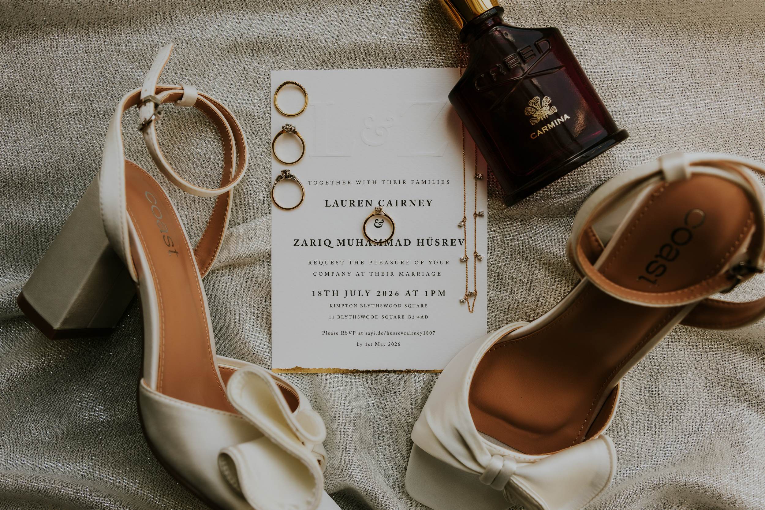Flatlay of ivory bridal shoes, three wedding rings on an invitation and a perfume bottle.