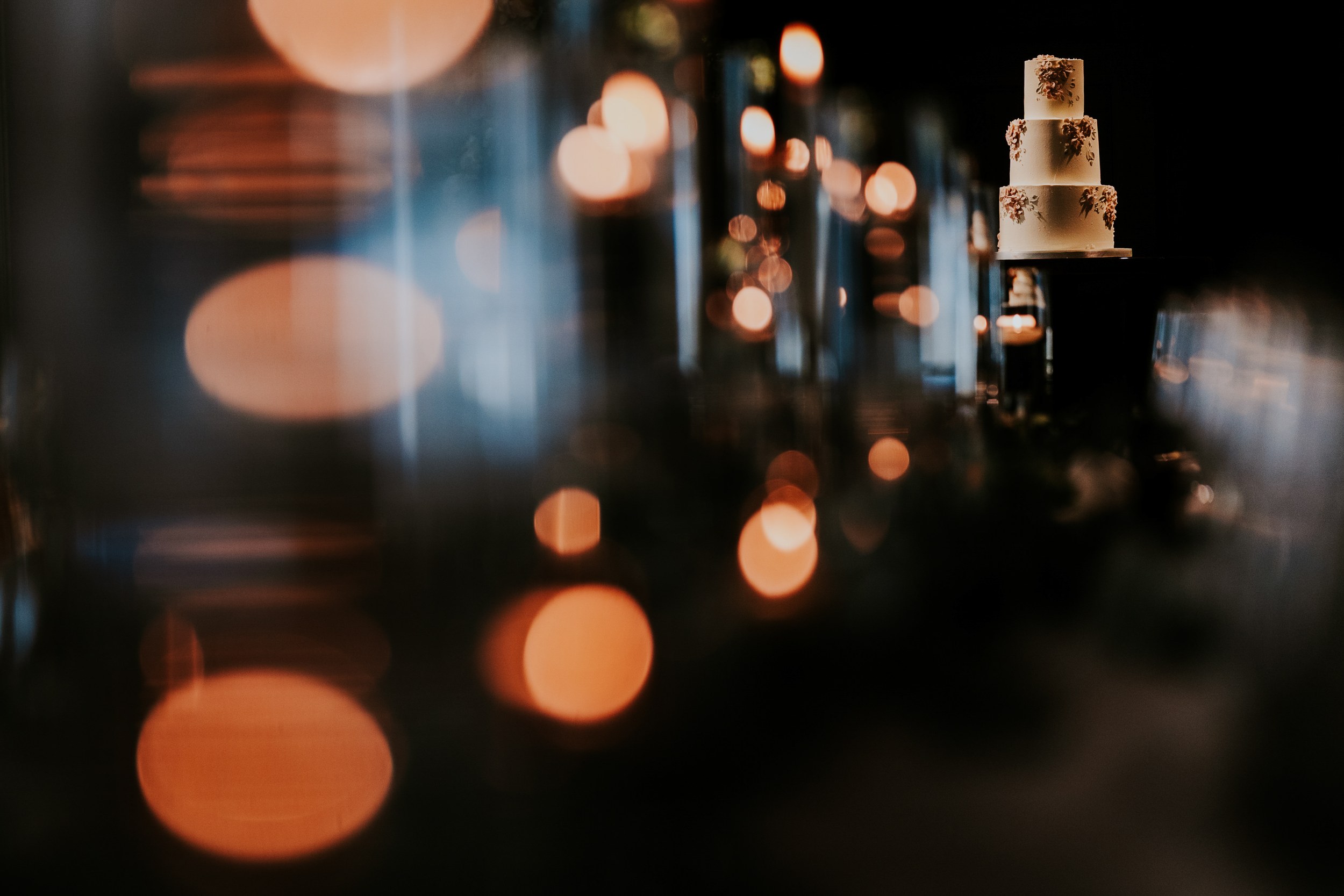 Four-tier white wedding cake with floral decorations on a dark stand, warm bokeh candle lights in the foreground.