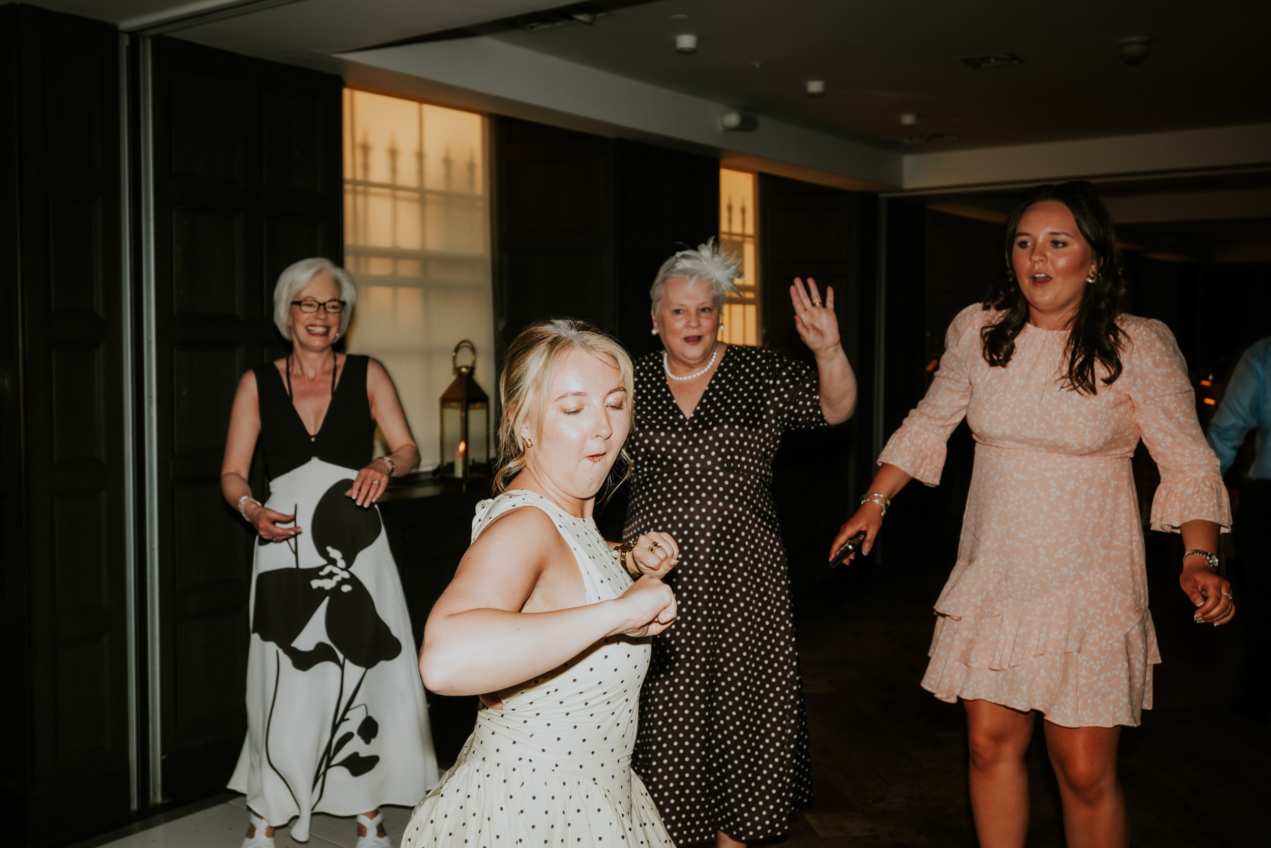 Four women dancing at a wedding reception indoors, one mid-dance in a pale polka-dot dress.