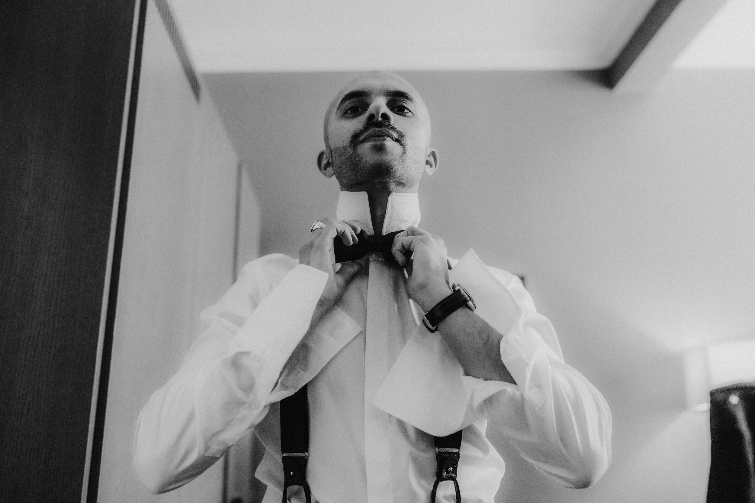Groom adjusting his bow tie in shirt and suspenders, low-angle black-and-white portrait.