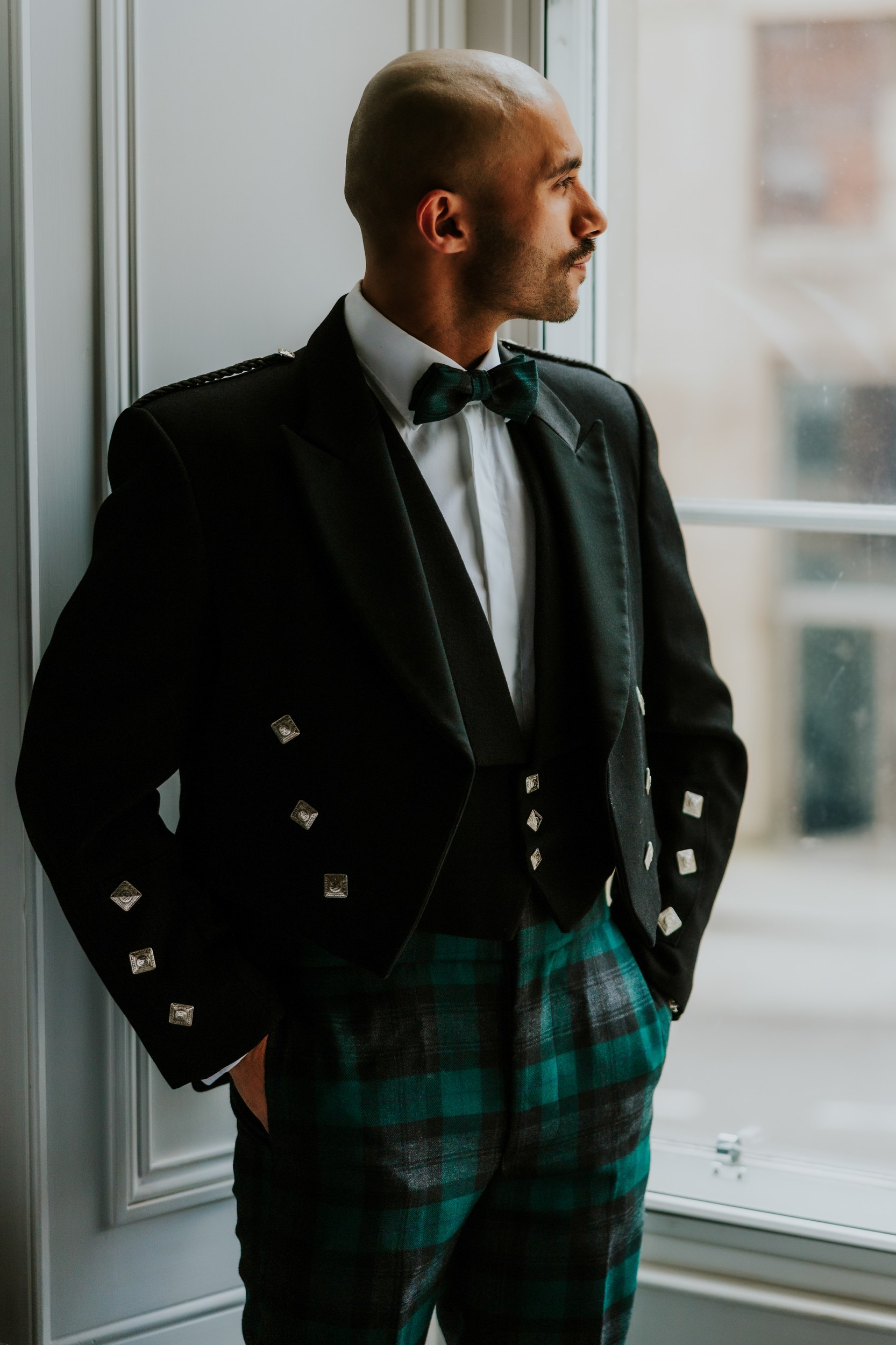 Groom in black doublet jacket and green tartan trousers looking out of a window.