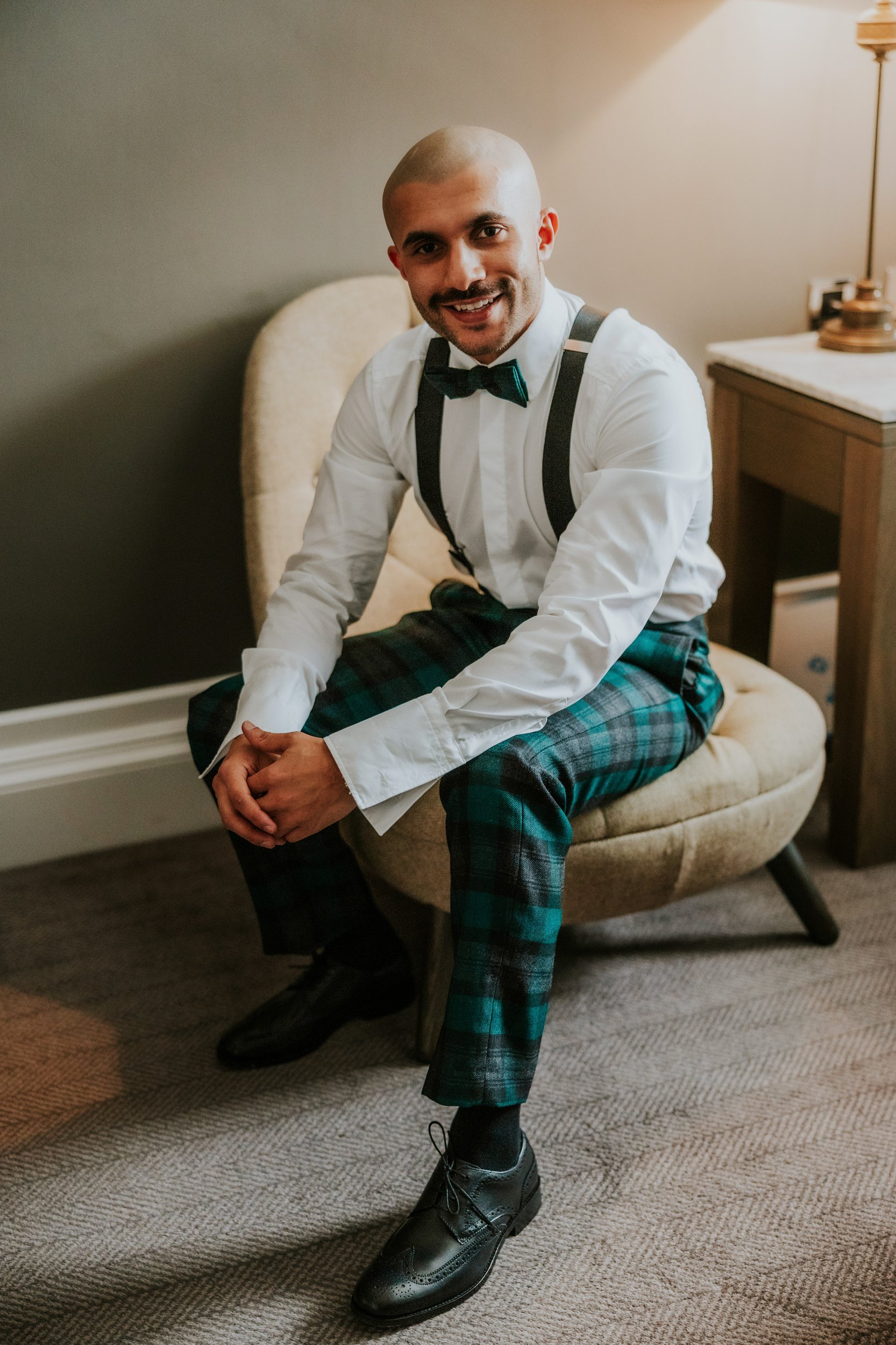 Groom seated in a hotel room wearing bow tie, suspenders and green tartan trousers, smiling.