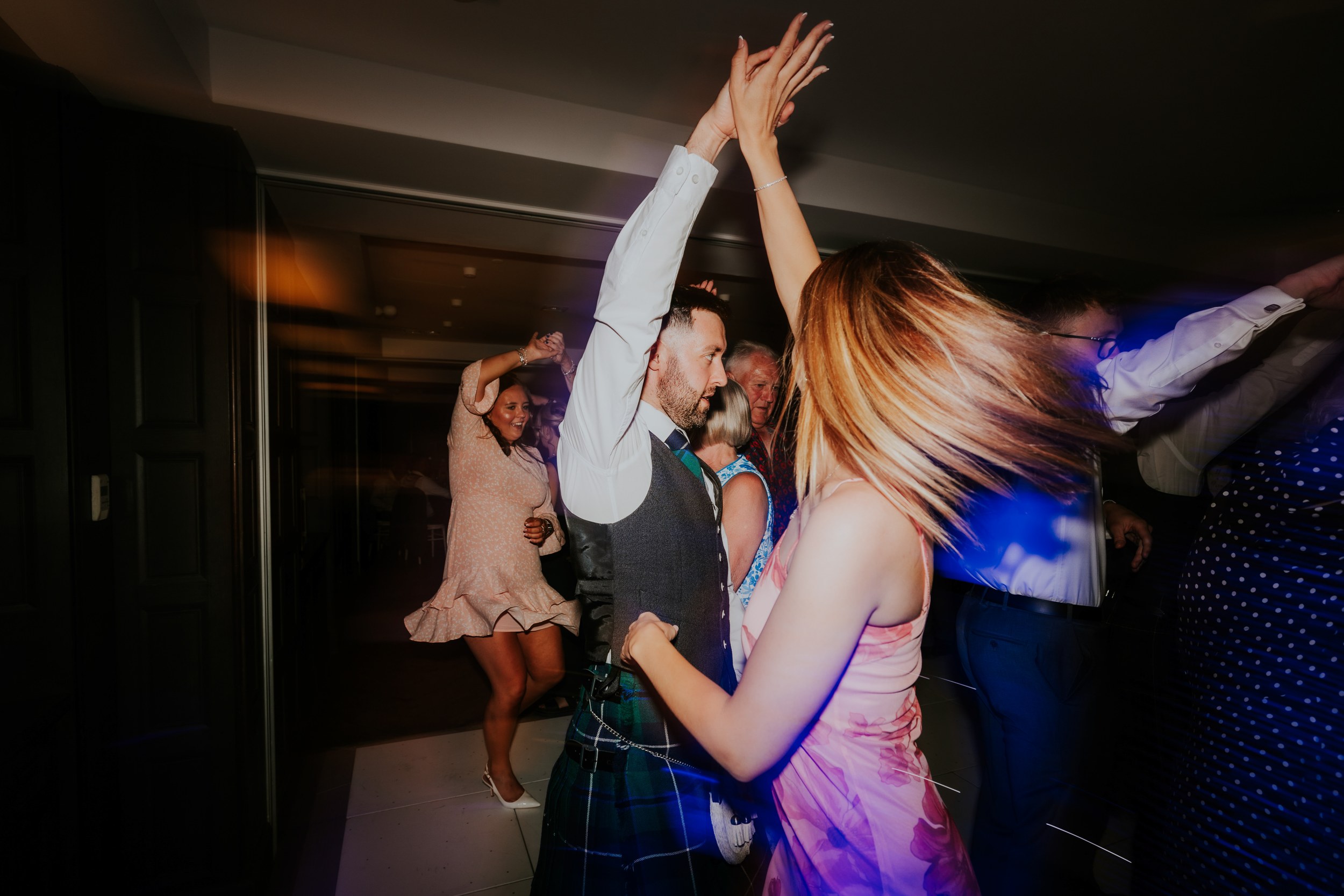 Guests dancing at a wedding reception; a man in a kilt spins a woman in a pink dress with hands raised.