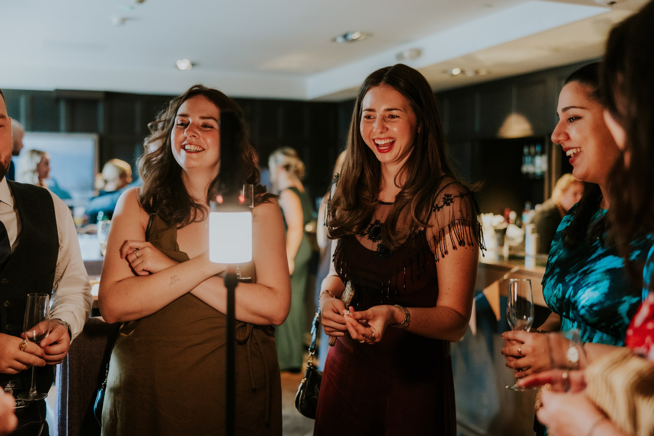 Guests laughing around a lit table lamp while holding champagne flutes in a dim reception lounge.