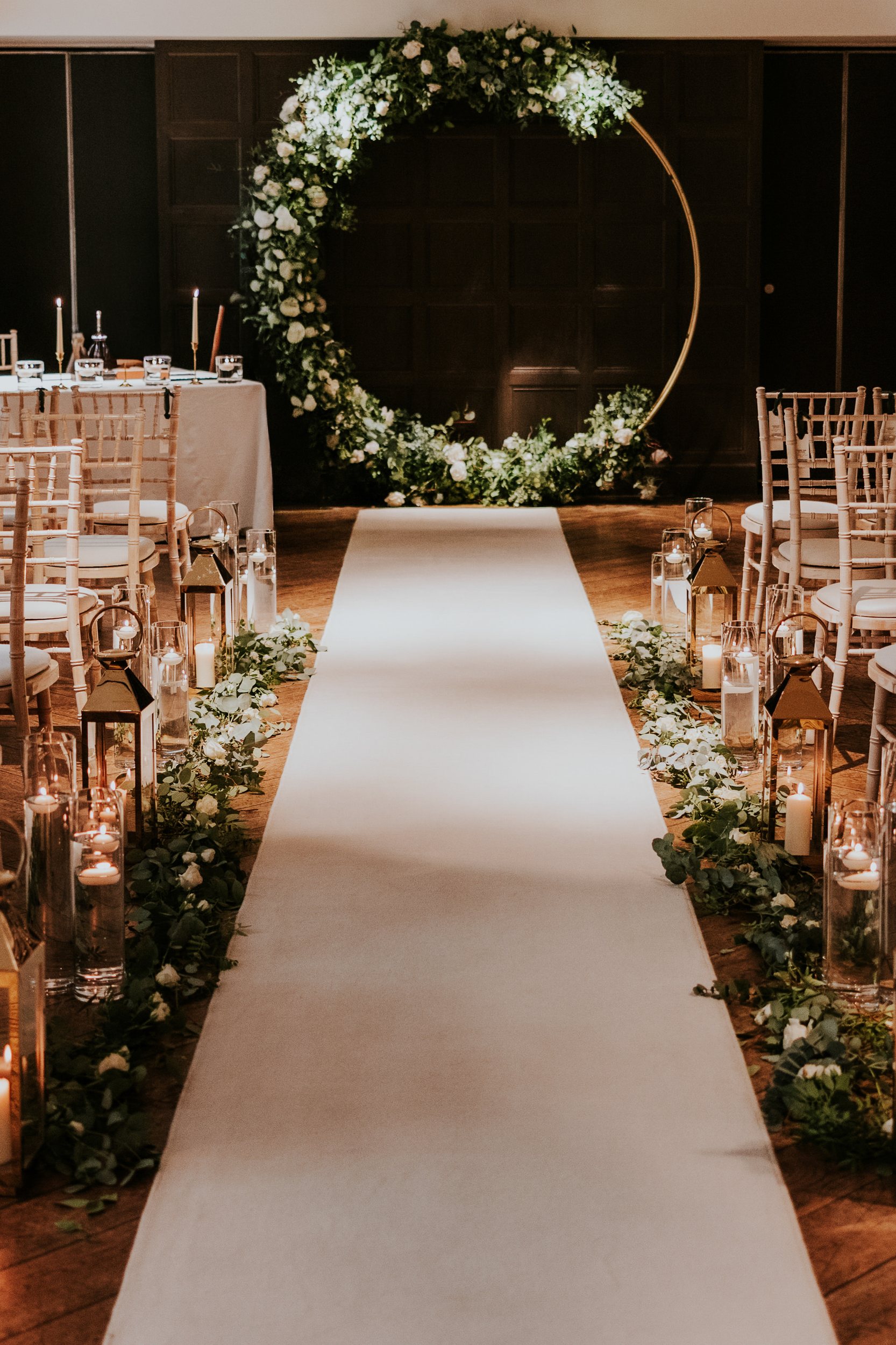 Indoor wedding aisle with white runner, candles and lanterns, foliage-lined edges leading to a circular floral arch.