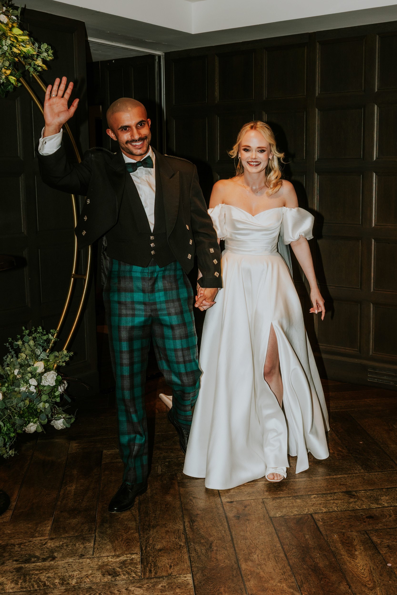 Lauren and Zariq enter holding hands — groom waves in jacket and green tartan trousers, bride in off-shoulder satin gown.