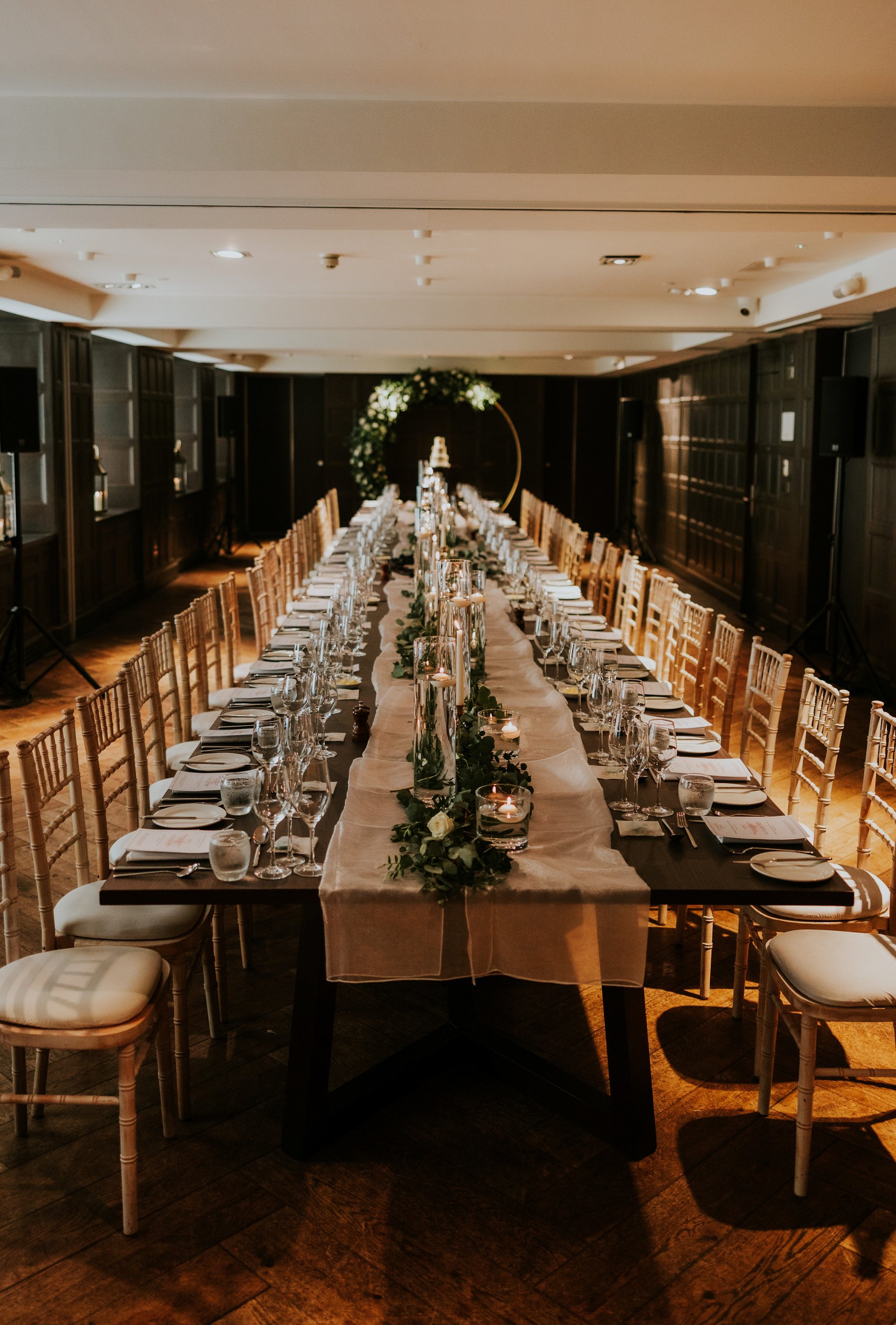 Long wedding reception table with candles, glassware and greenery in a wood‑panelled room and a circular floral arch at the end.