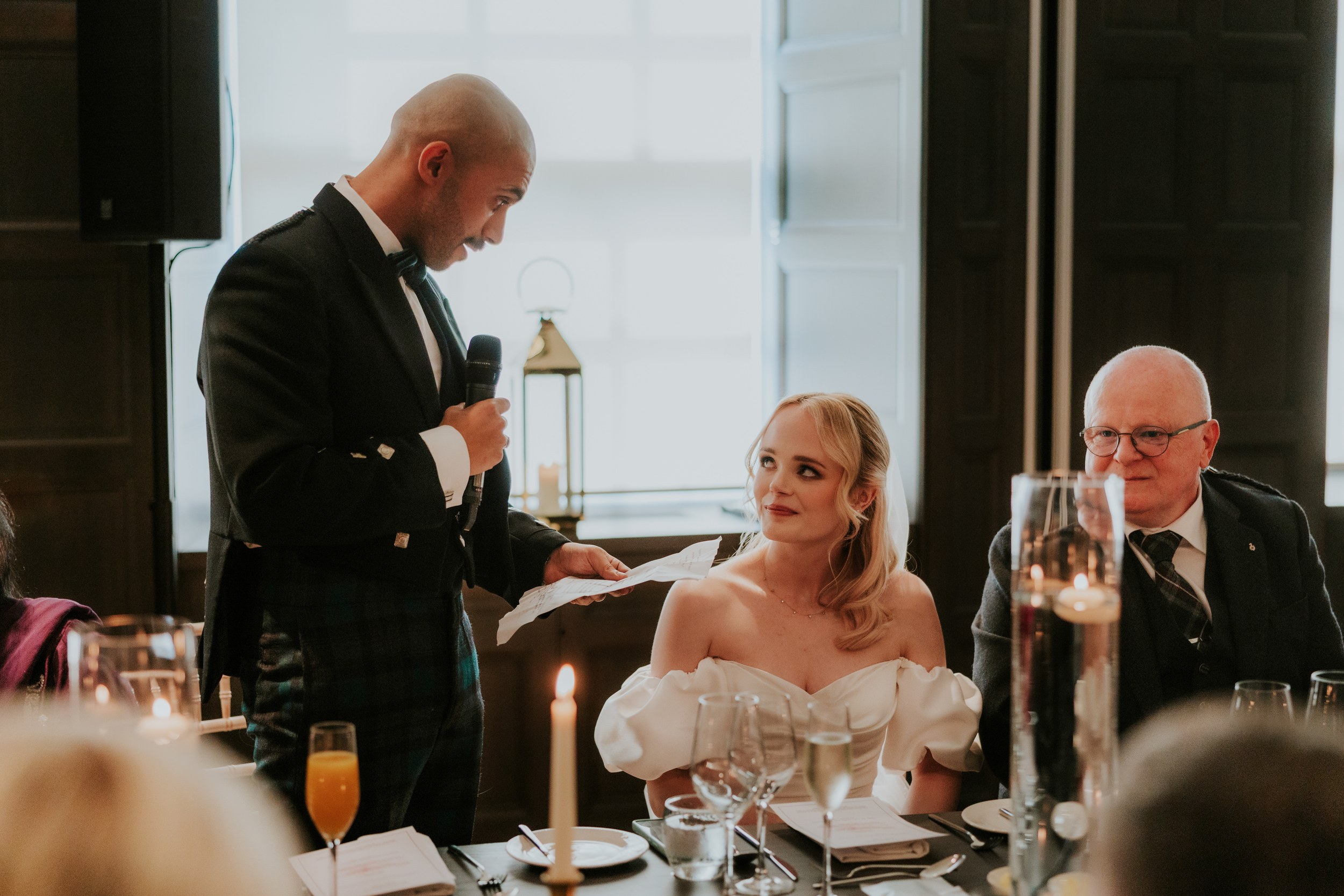 Man in formal attire holding a microphone reads from notes as a bride in an off-the-shoulder dress listens at the reception table.