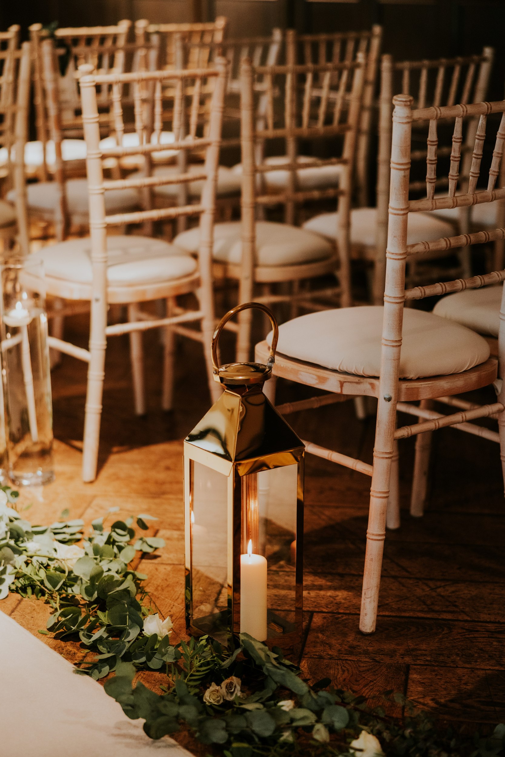 Pale wooden Chiavari chairs with cushions, aisle greenery and a gold lantern with a lit candle on a wooden floor.