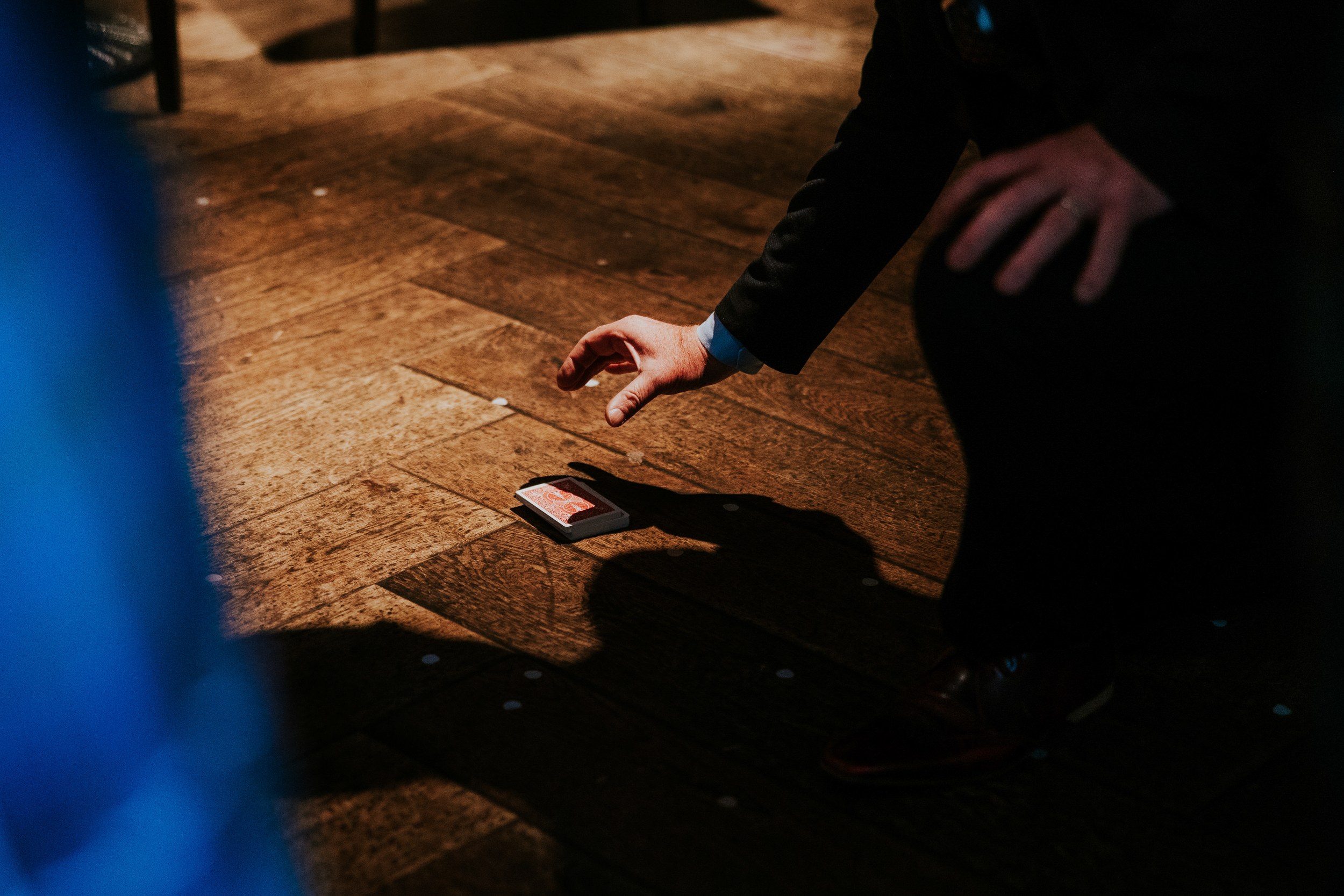 Person's hand in a suit reaches for a deck of playing cards on a wooden floor with confetti.