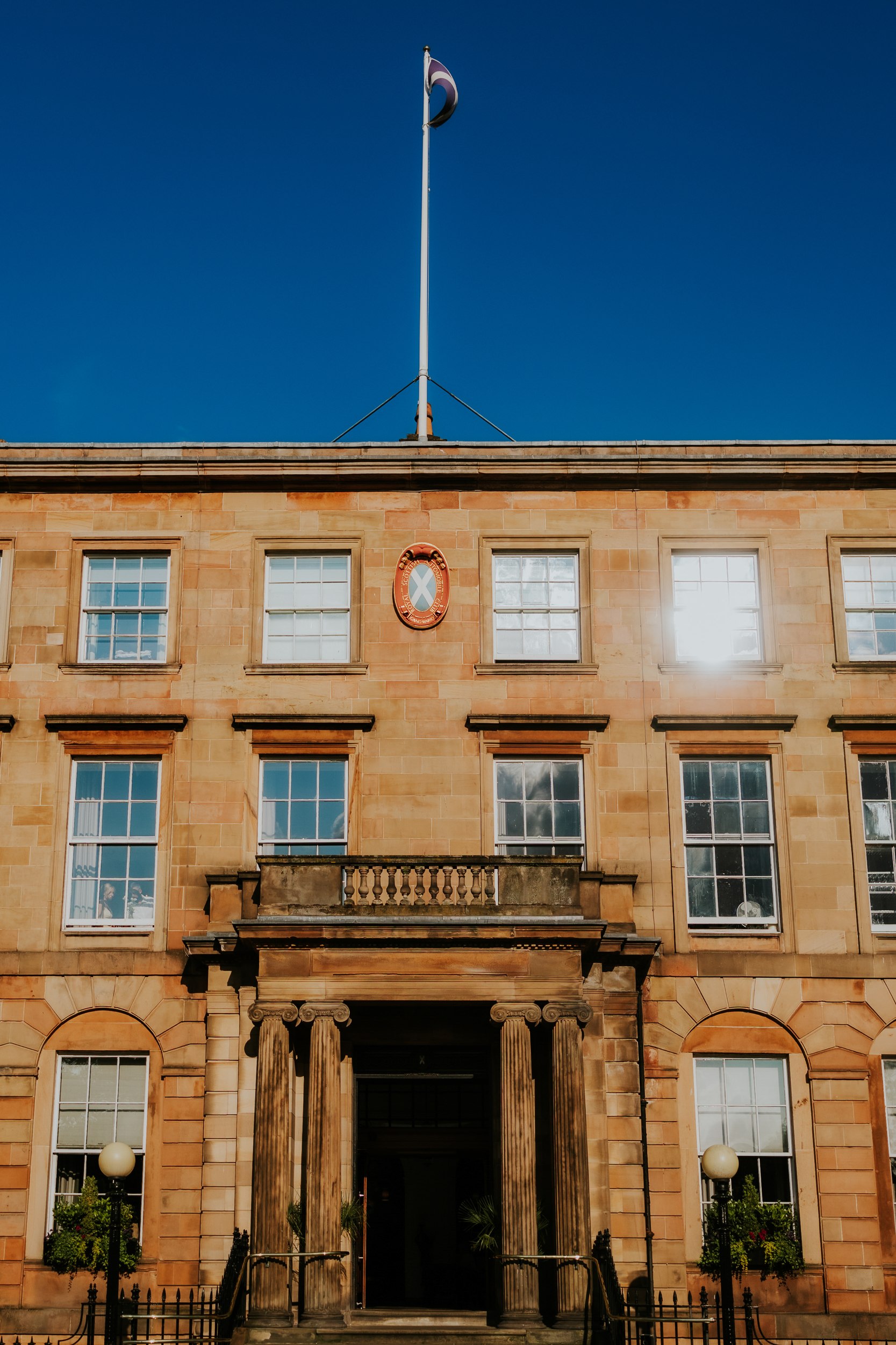 Sandstone façade with central balcony, columned entrance, sash windows and a flagpole against a clear blue sky.