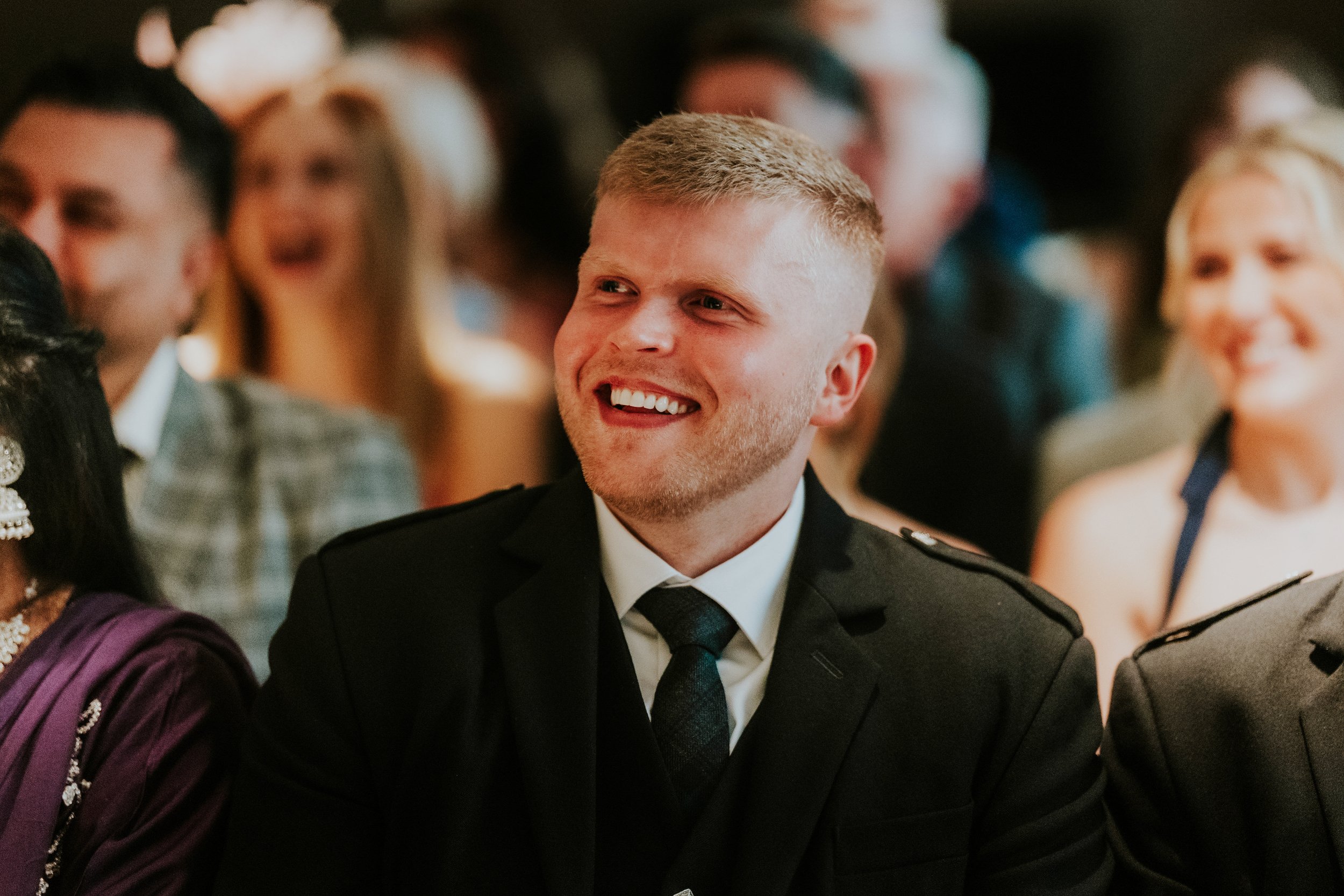 Smiling male wedding guest in dark suit and tie, seated with blurred guests behind him.