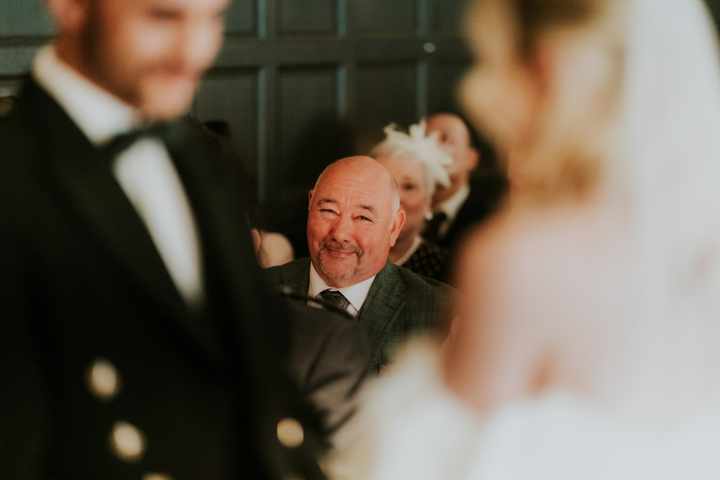 Smiling seated guest framed between the blurred bride and groom at a wedding.