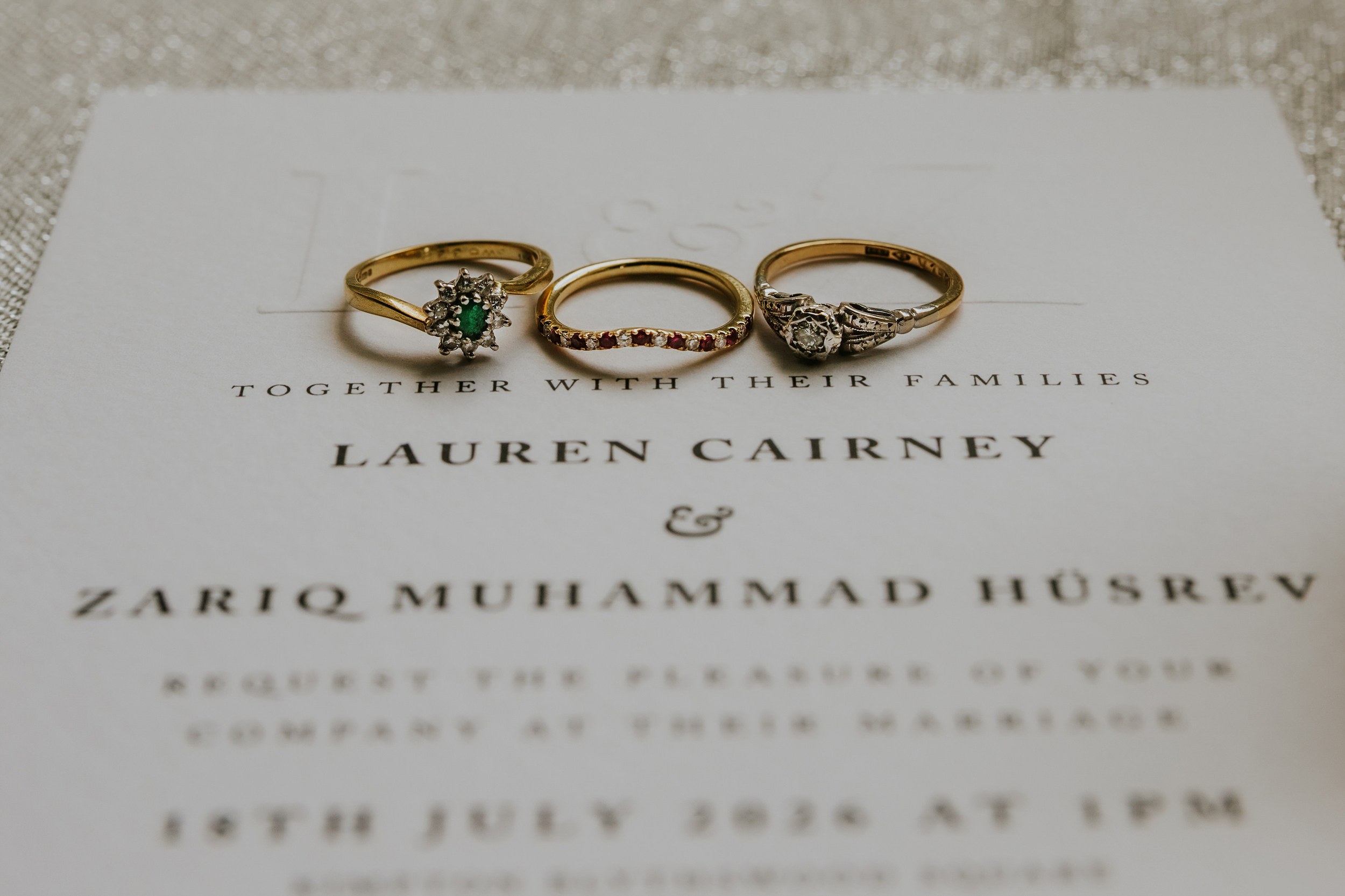 Three gold rings on a wedding invitation showing 'Lauren Cairney' and 'Zariq Muhammad Hüsrev'.