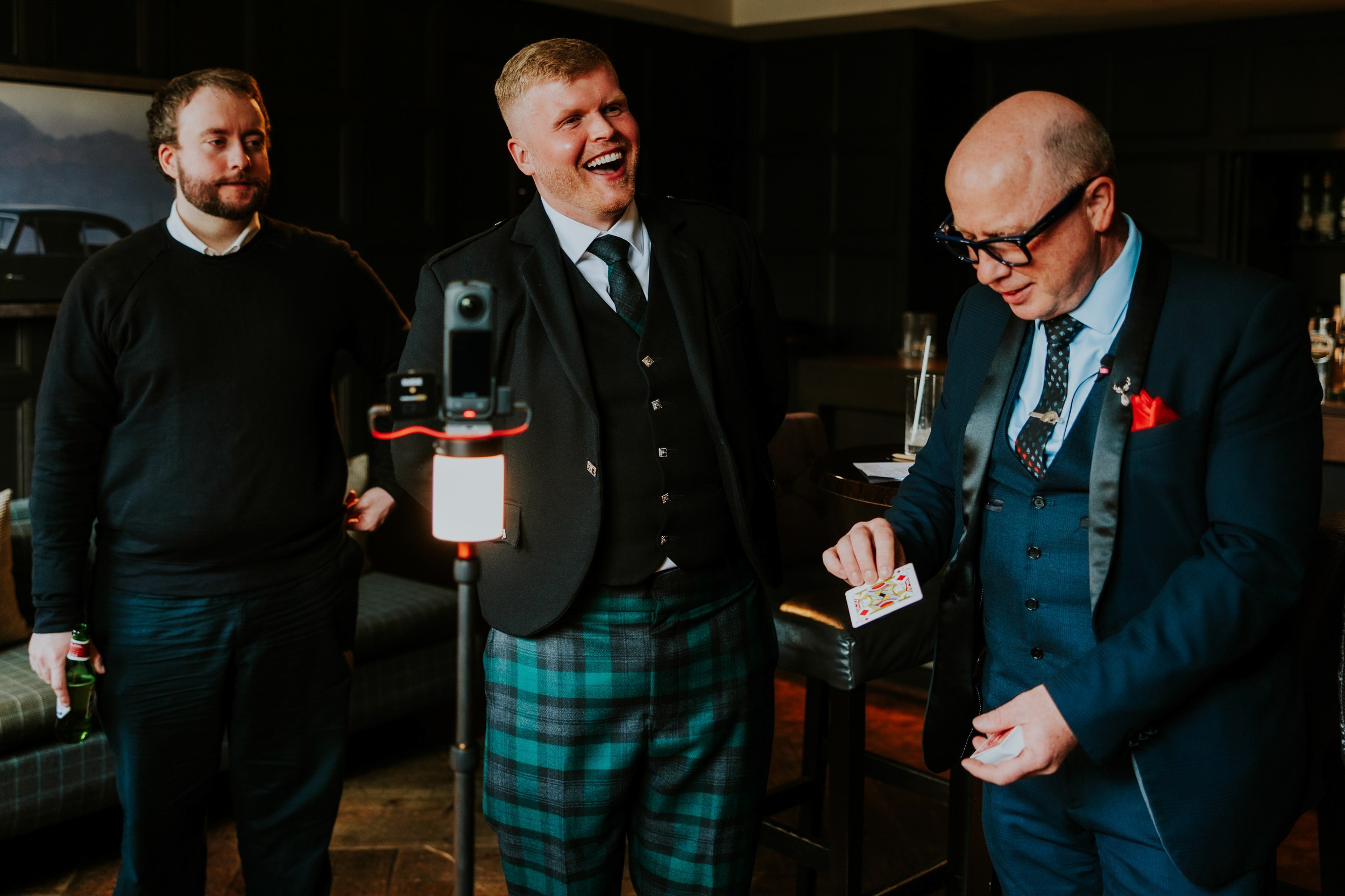 Three men in a hotel lounge; groom in green tartan kilt laughs as a guest performs a card trick by a tripod camera.