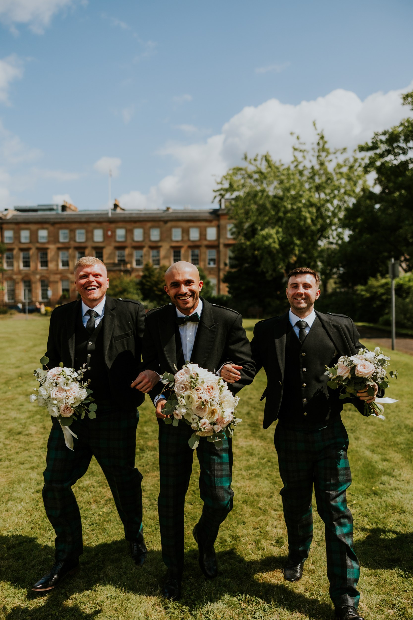 Three smiling groomsmen in kilts holding bouquets walking on a lawn in front of a sandstone building.