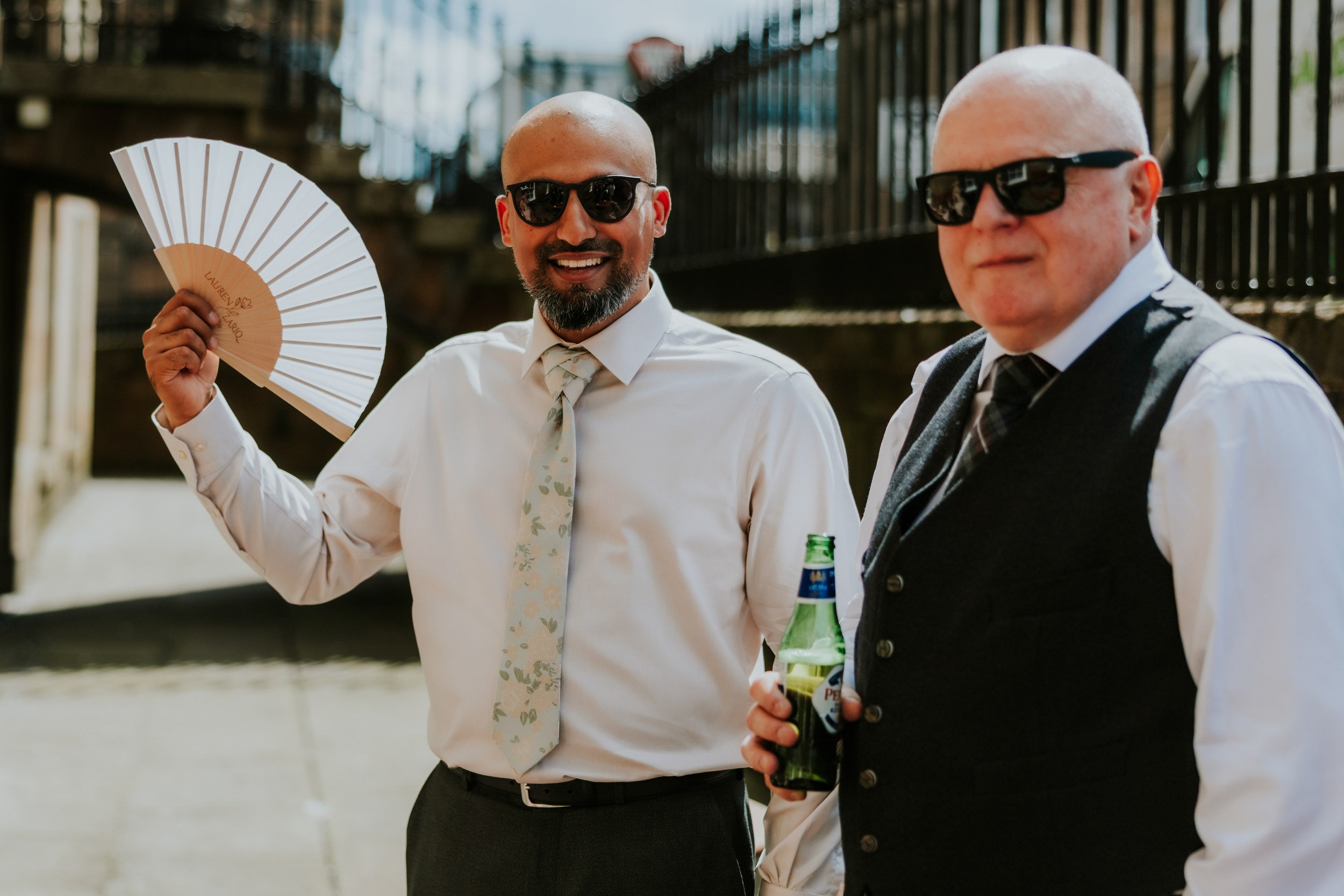Two male wedding guests in shirts and sunglasses, one holding a personalised fan, the other holding a beer bottle.