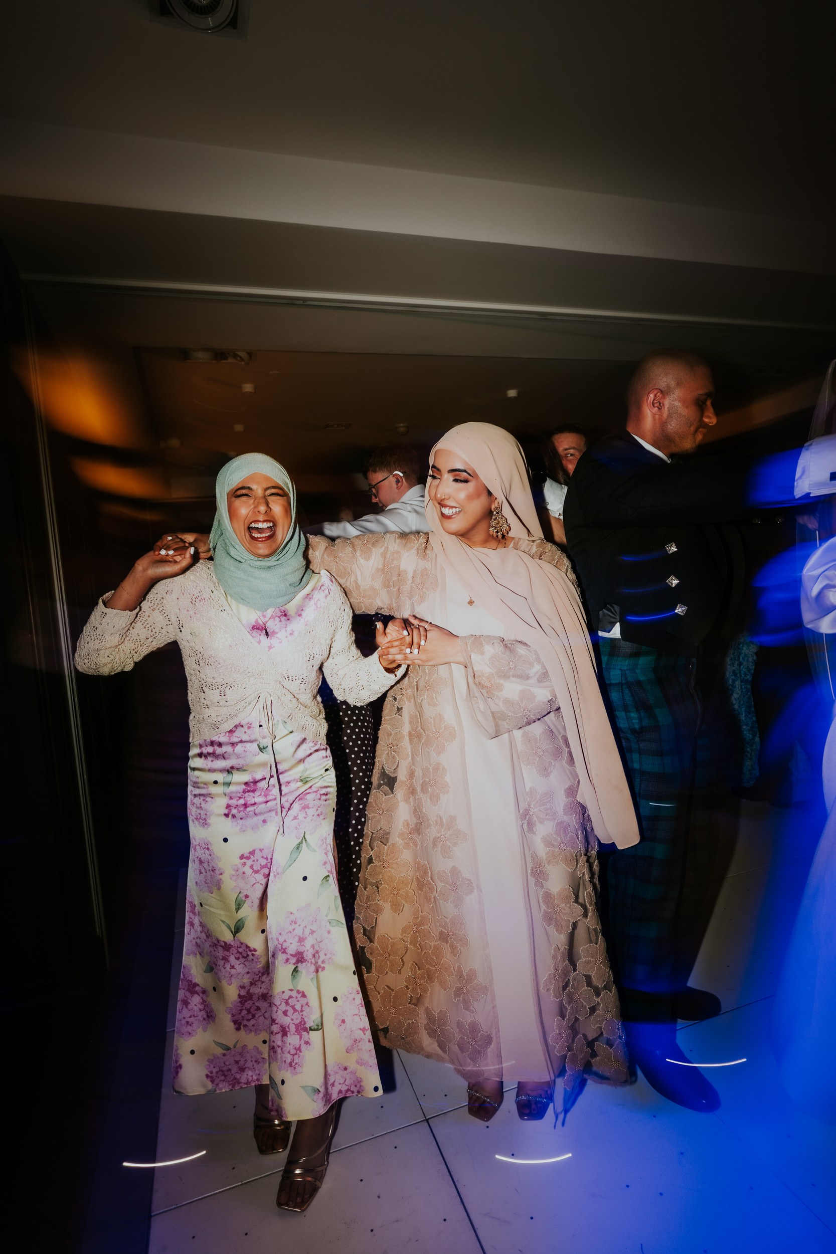 Two women in hijabs smiling and holding hands while dancing at a wedding reception, with guests and a man in a kilt behind them.