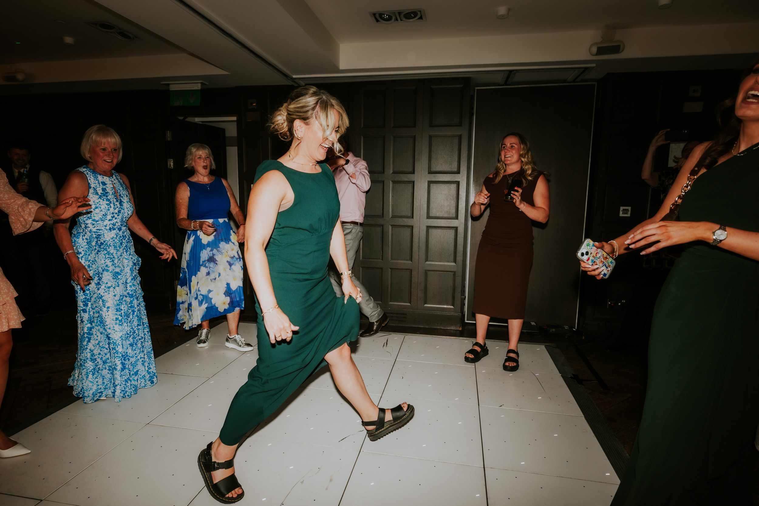 Wedding guests dancing on an indoor white dance floor; woman in a green dress mid-step while others smile and hold drinks.
