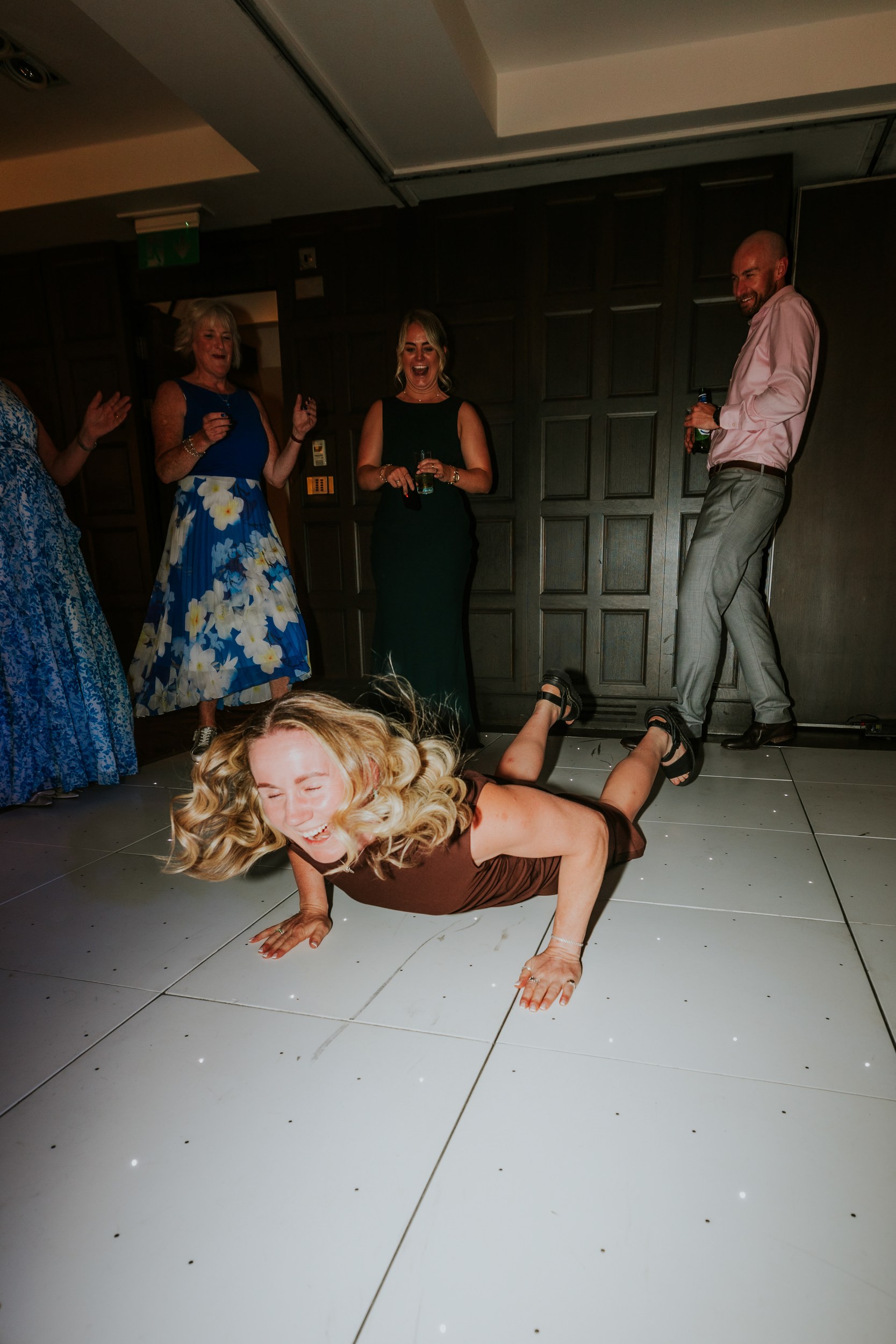Wedding guests on a dancefloor; a woman laughing on the floor while others stand behind her with drinks.
